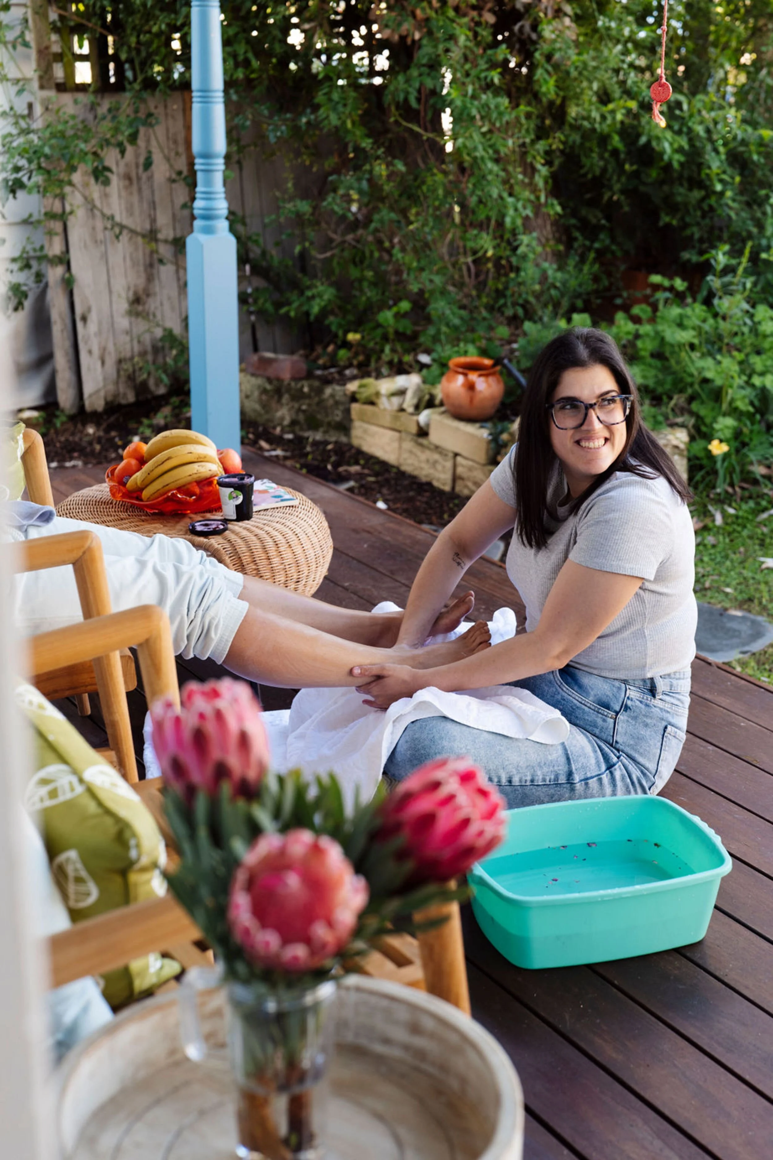 A postpartum doula with glasses sitting on a wooden deck, smiling, with a woman lying back with her legs on her lap. A table with fruit and a flower vase in the foreground, and green foliage and a brown pot in the background.