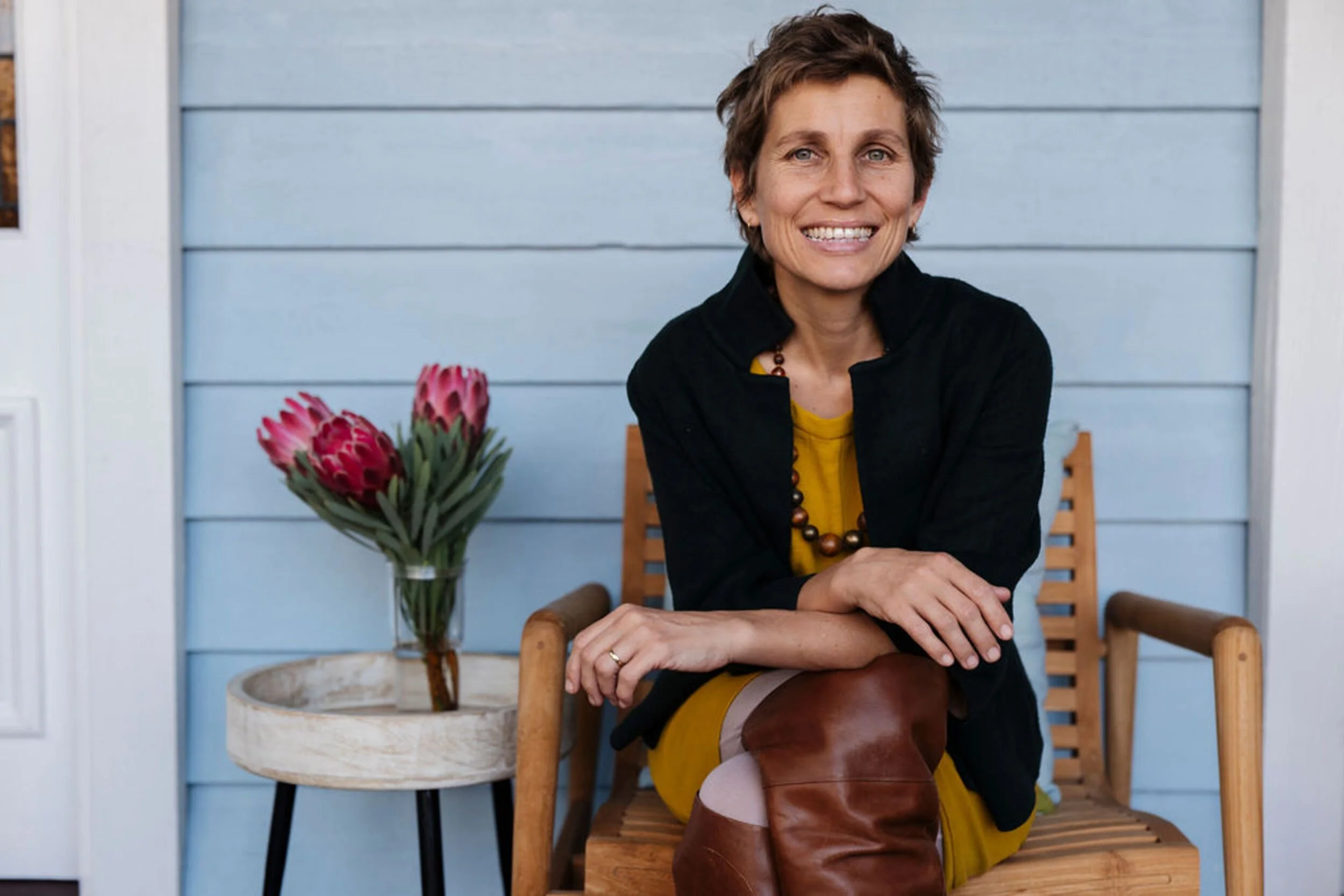 A woman with short brown hair, wearing a black jacket, yellow dress, and brown boots, sitting on a wooden chair, smiling at the camera, with a vase of pink protea flowers on a round table to her left, and a blue wall in the background.