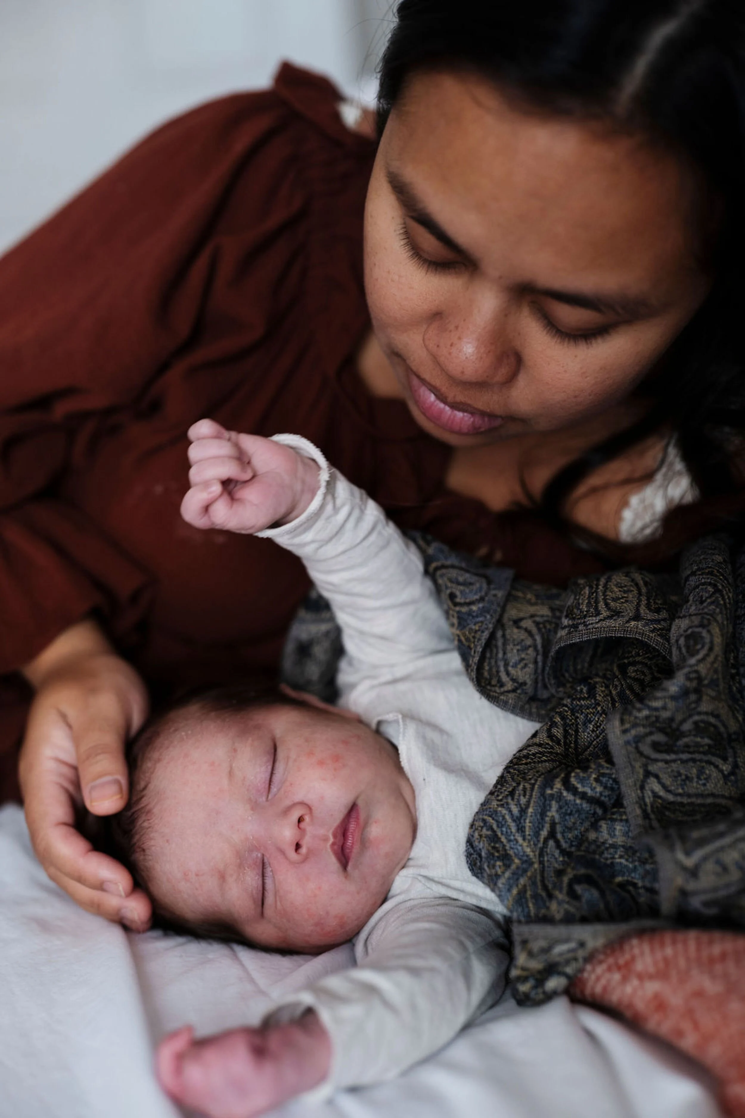 A woman with dark hair and medium skin tone gently holds a sleeping baby's head, who has light skin and dark hair, on a white sheet.