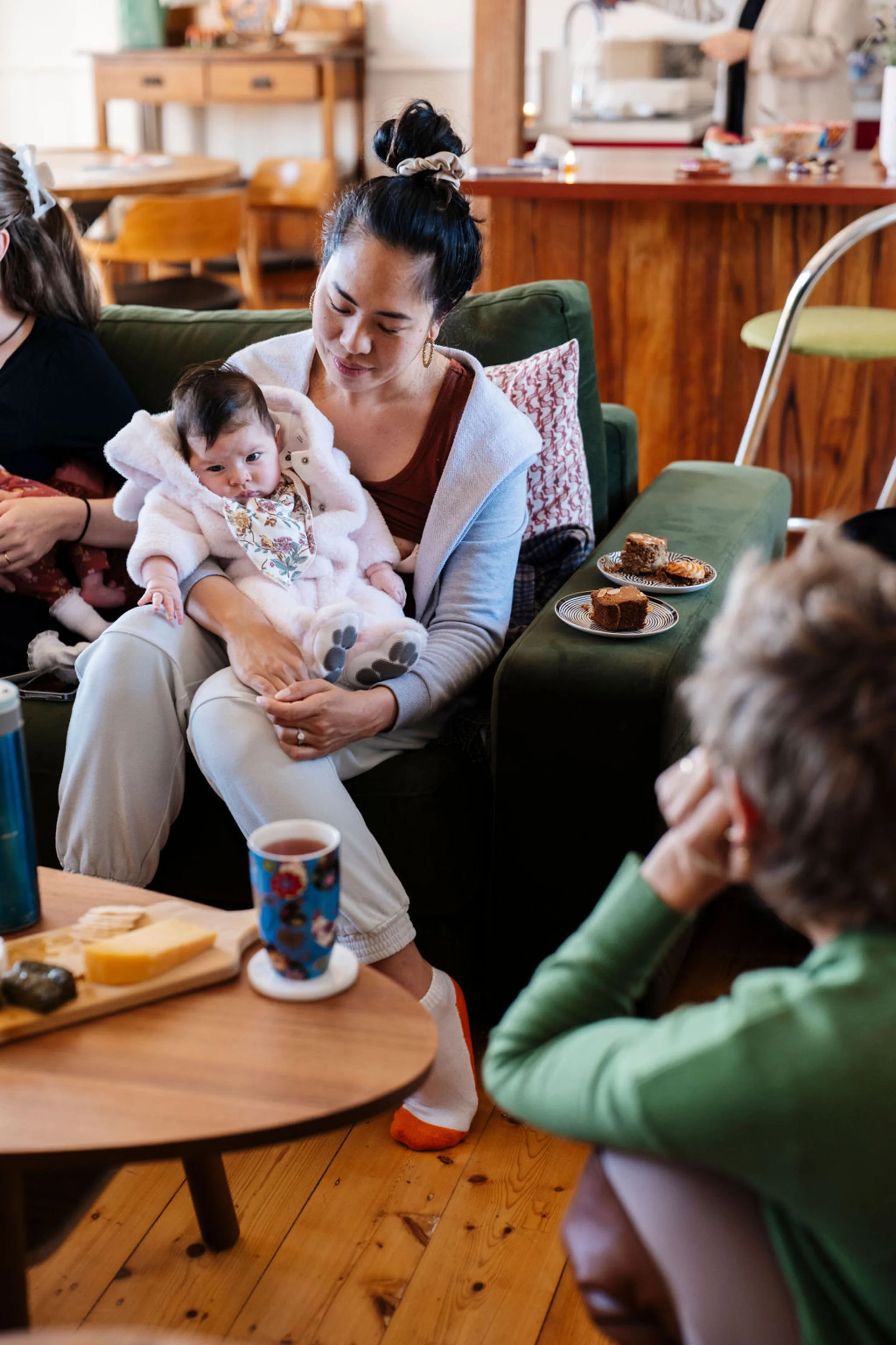 A mother holding her baby, and being support by a certified postpartum doula, sitting on a green sofa holding a baby in a cozy indoor setting.There are plates of cake on a table nearby, along with a cup and some snacks.