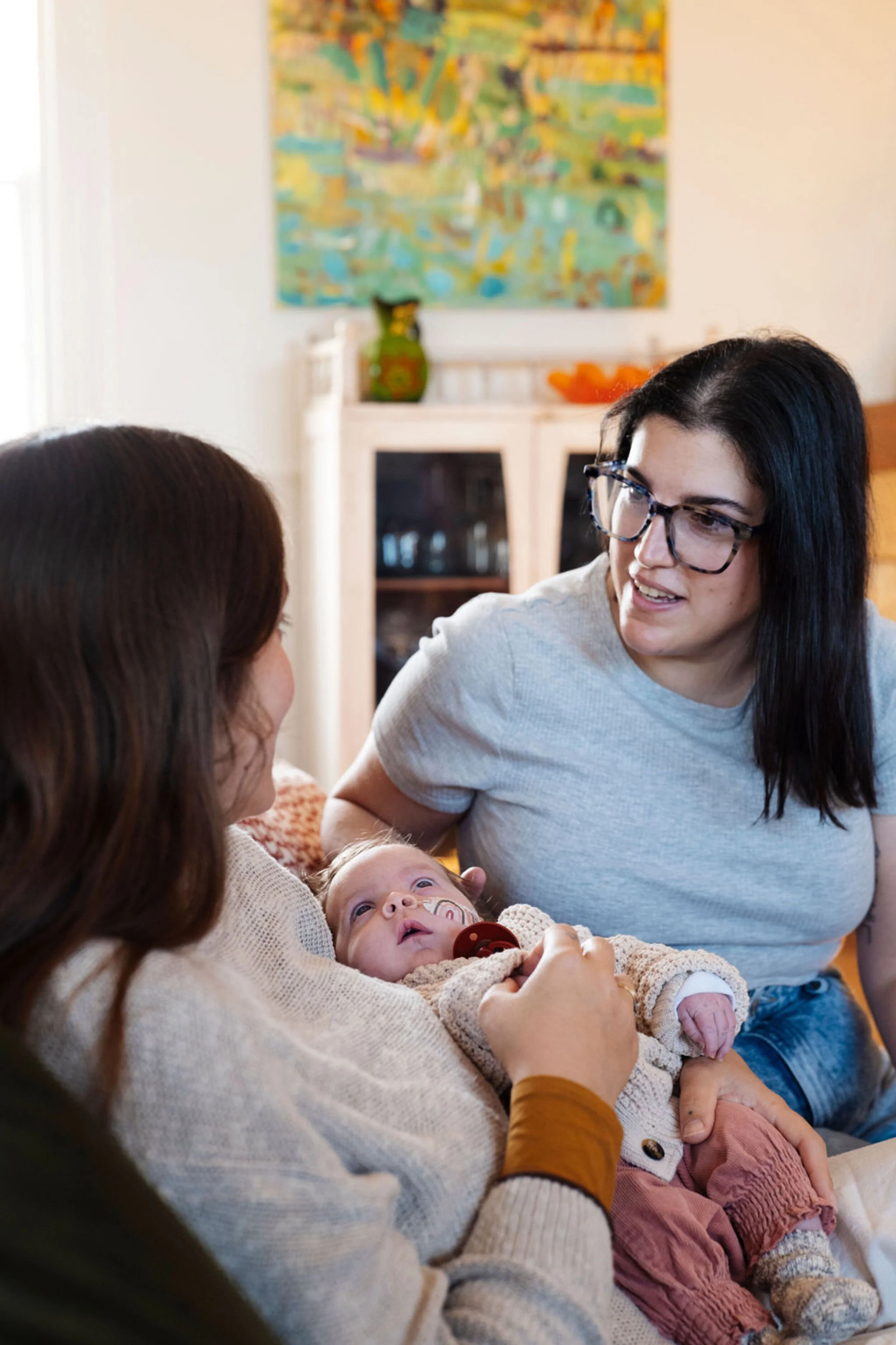 A mother with glasses holding a baby on her lap, with a postpartum doula talking to her, in a cozy room with a painting on the wall and a cabinet in the background.