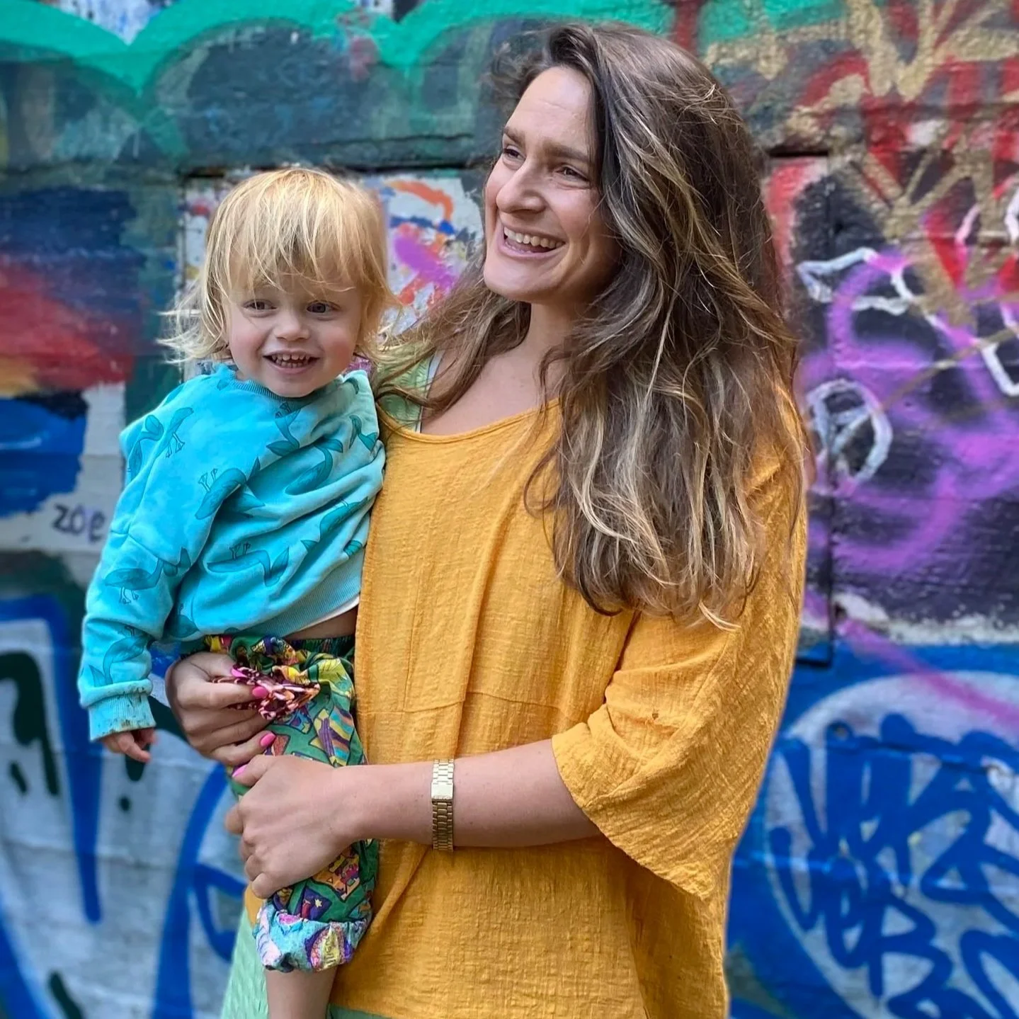 A woman with long wavy brown hair smiling while holding a young child with blond hair and a big smile, standing in front of a colorful graffiti wall. The woman is wearing a yellow top and a silver watch, and the child is dressed in a blue hoodie and colorful pants.