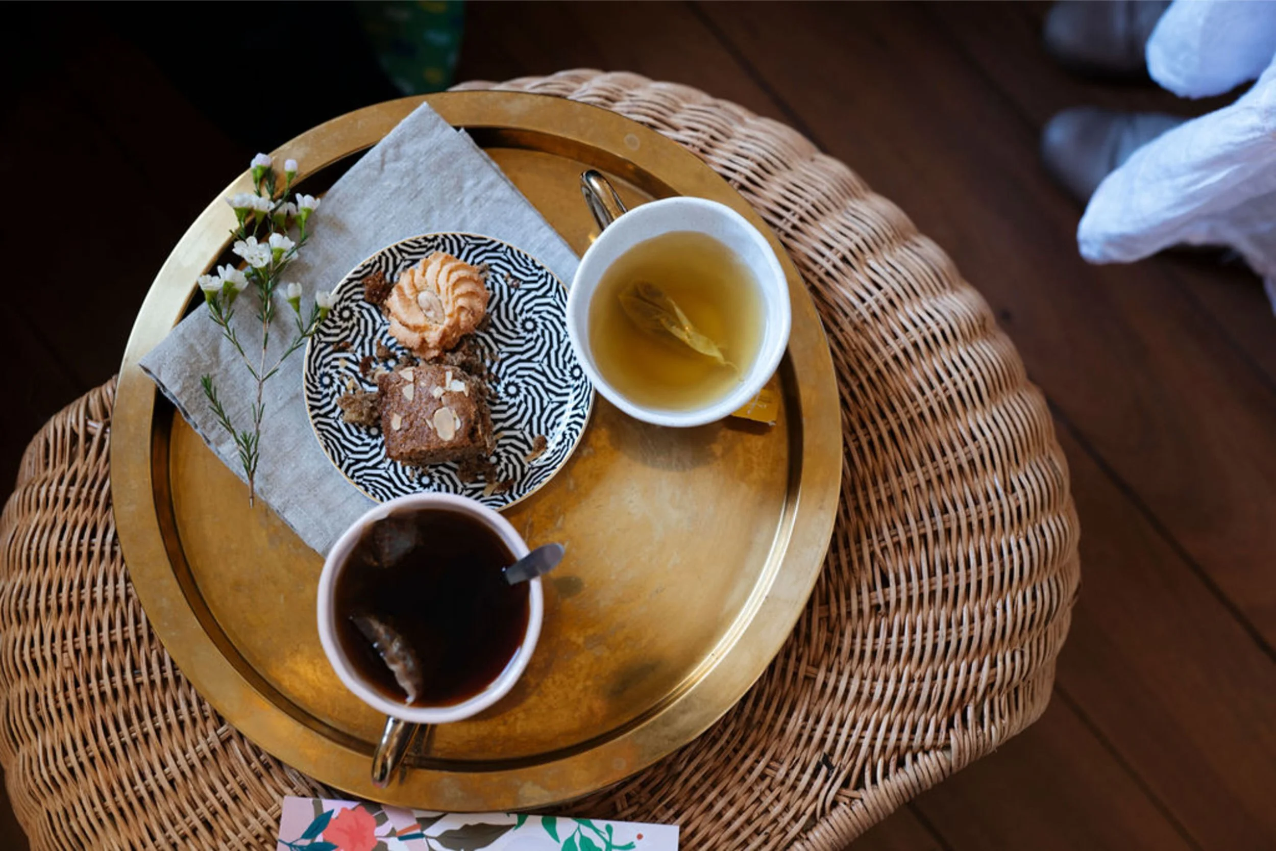 A round golden tray with a cup of tea, glasses of coffee, and a plate of assorted cookies, resting on a wicker table with a white napkin, flowers, and a decorative card.