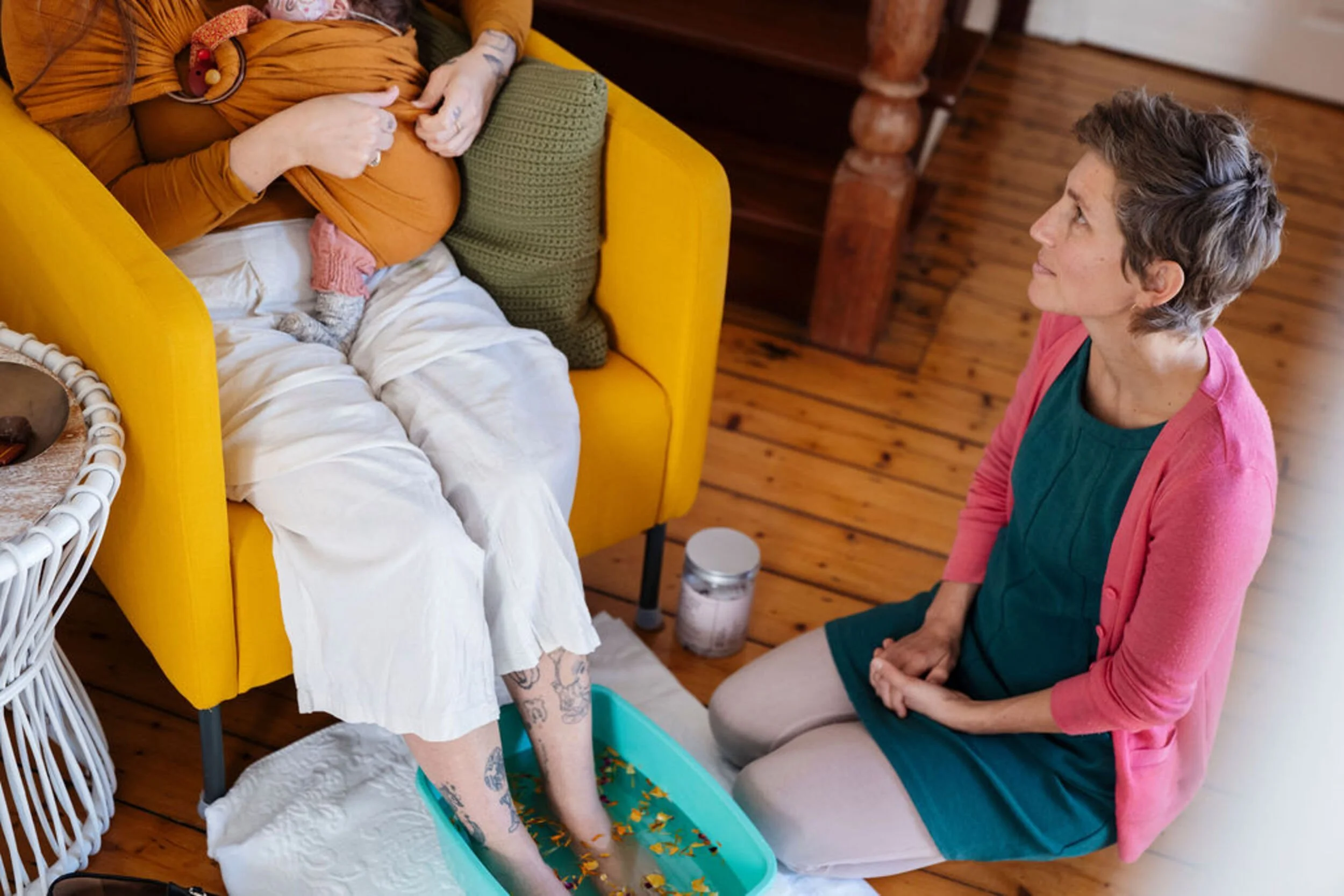 A certified postpartum professional with short hair sitting on the floor, looking up at a mother with a baby in a carrier in a yellow armchair with their legs in a foot spa filled with water.
