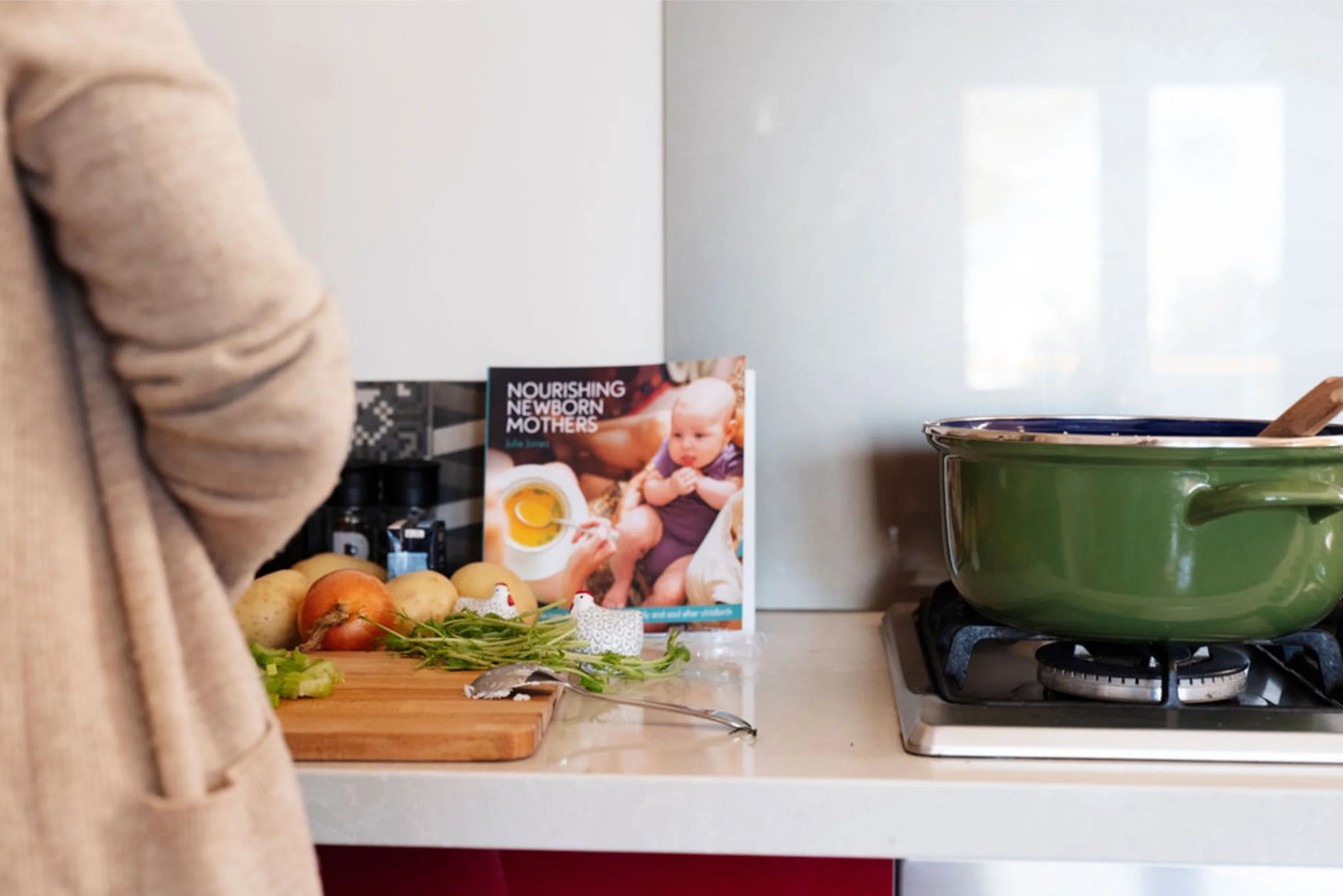 A certified doula cooking at a kitchen counter with a green pot on a stove, a wooden cutting board with chopped vegetables, onions, and herbs, and a cookbook titled 'Nourishing Newborn Mothers' by Julia Jones