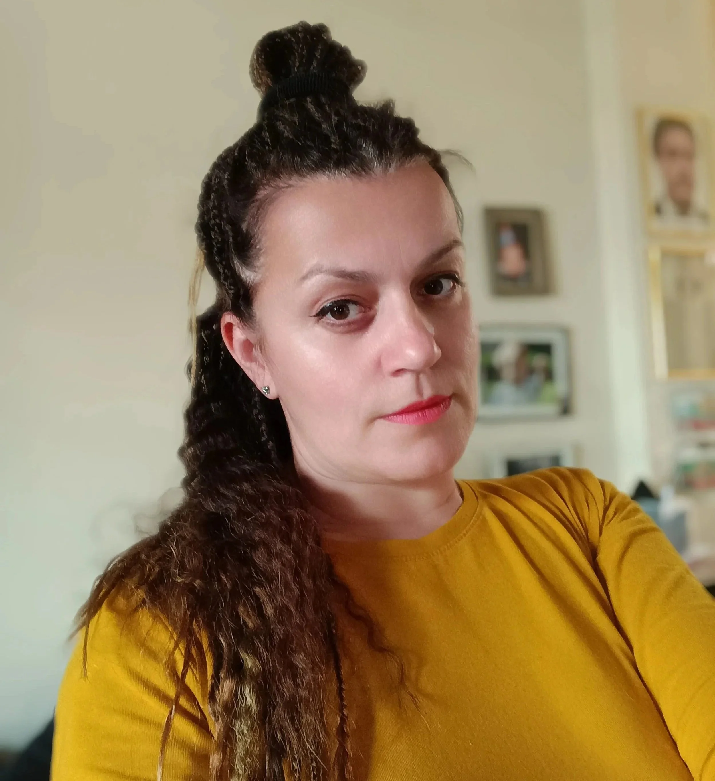 A woman with dark braided hair and a brown fur shoulder draped over a mustard yellow top, standing indoors in front of a beige wall with framed photos.