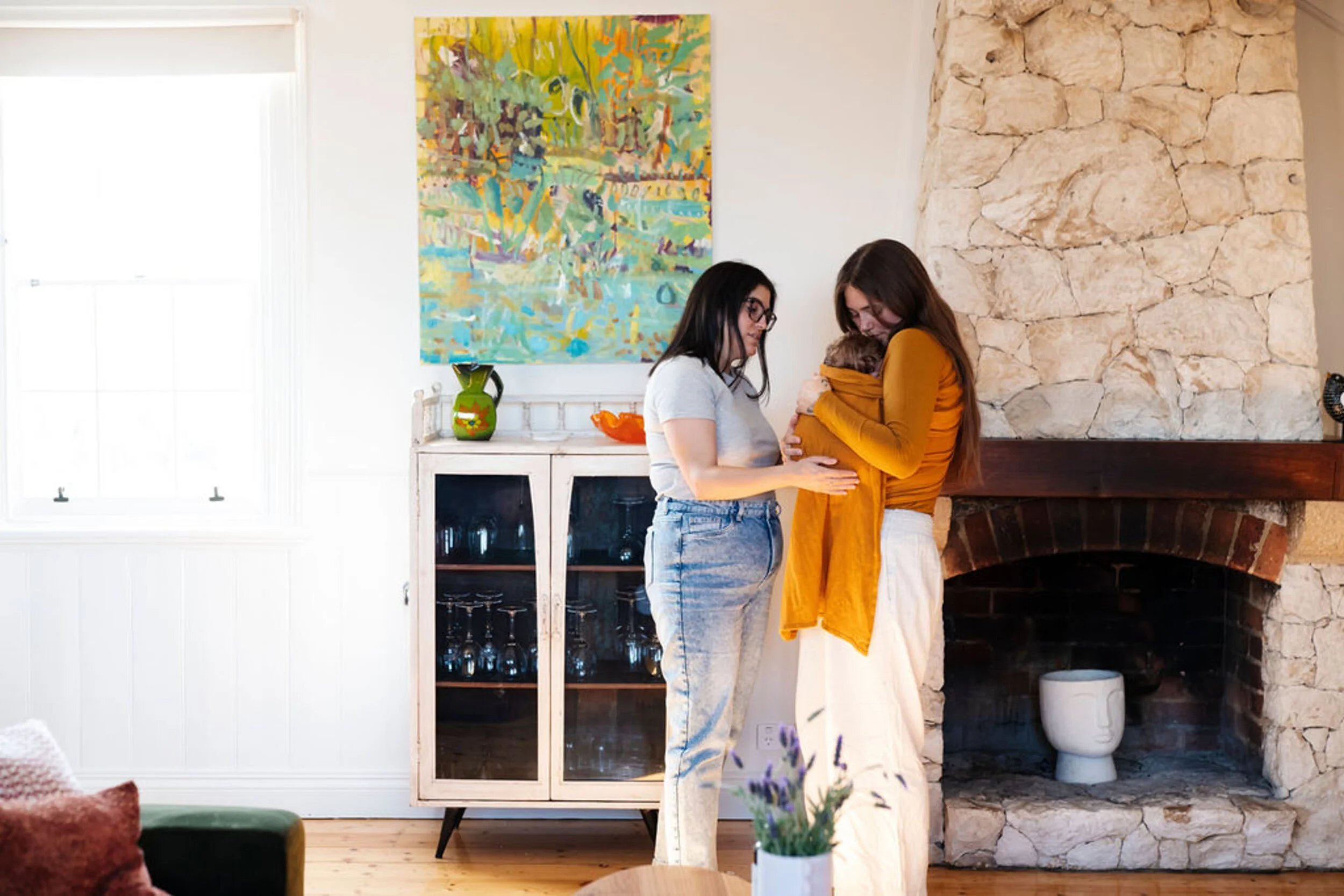 A postpartum professional supporting a mother with her baby in a carrier. Behind them is a white cabinet with glass doors and a colourful abstract painting on the wall.