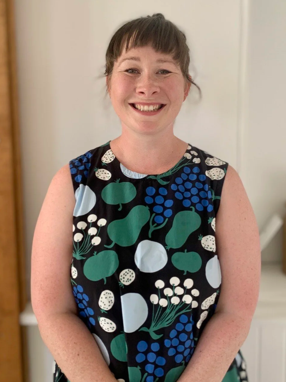 A woman with short brown hair smiling, wearing a sleeveless dress with a fruit and flower pattern, standing indoors against a neutral background.