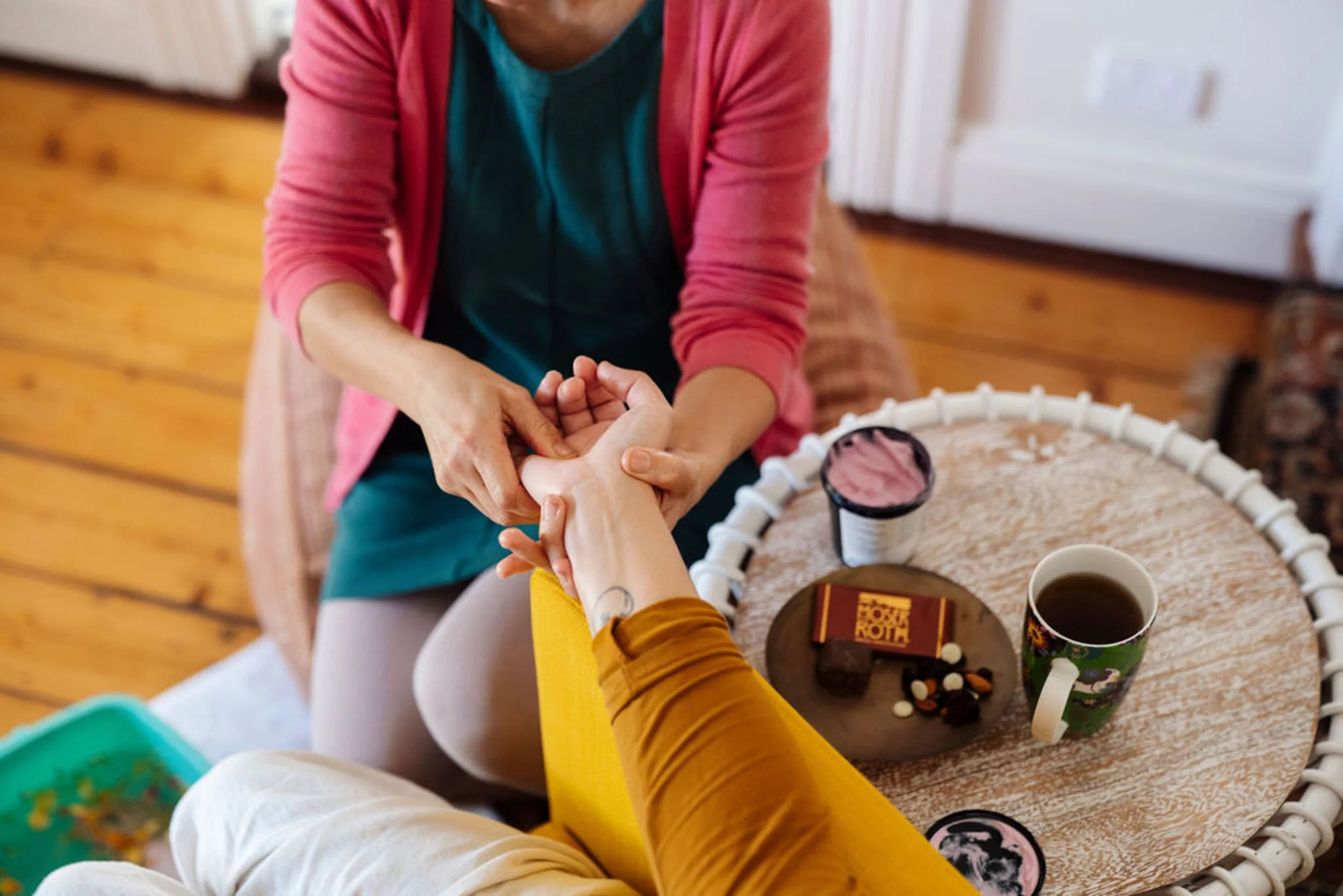 Certified postpartum doula with teal shirt and pink cardigan giving a hand massage to a new mother, with tea and chocolate on the table.