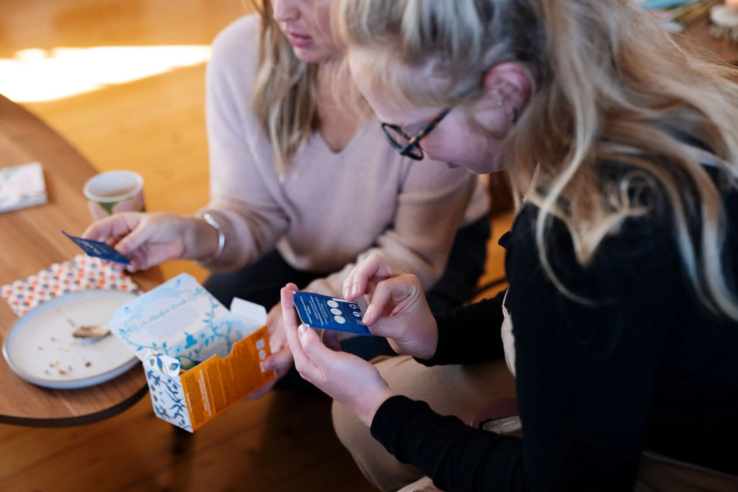 A mother and a postpartum doula sitting at a wooden table choosing from a box of tea, with a partially eaten plate of food and a small cup on the table.