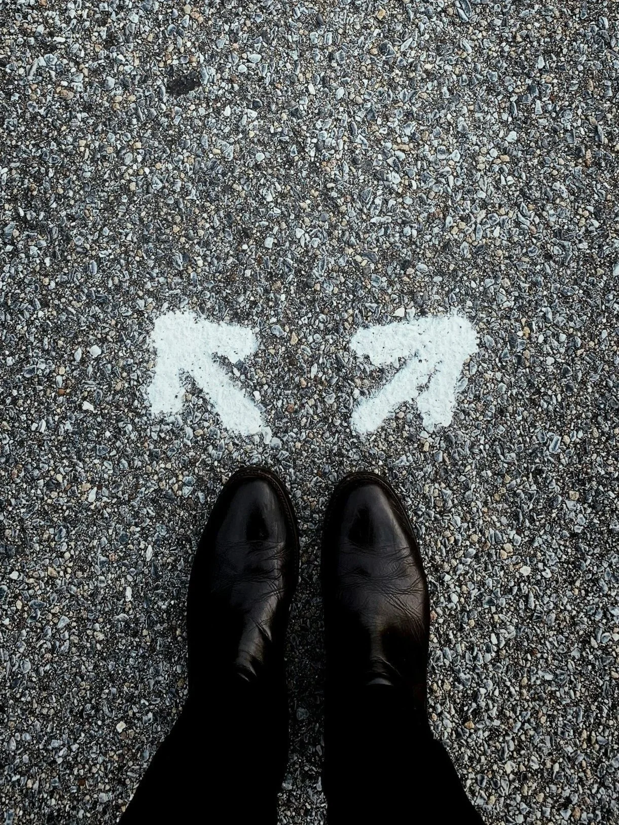 A person's black shoes and black pants standing on gravel ground with white painted arrows pointing left and right.