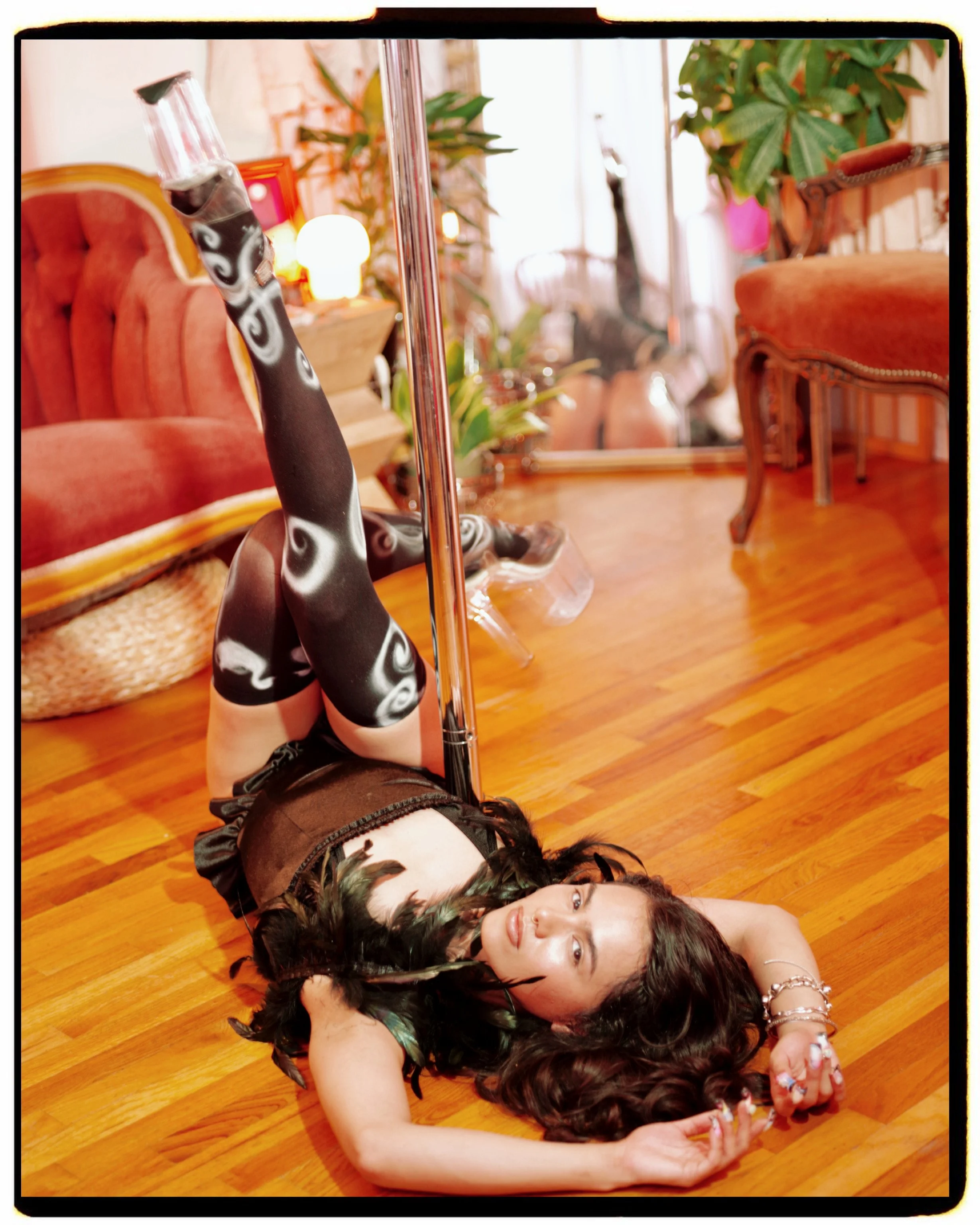 model and pole dancer Frankie Alvarez poses on the ground next to a pole wearing black thigh high tights airbrushed with white swirly accents and a black feathered vest top