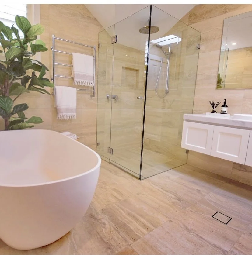 Modern bathroom with a freestanding bathtub, glass-enclosed shower, white vanity with a mirror, towel rack with towels, a potted plant, and a soap dispenser on the vanity, all with beige tiled walls and floor.