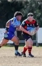 Two rugby players competing for the ball on the field, one in blue and one in red and black.