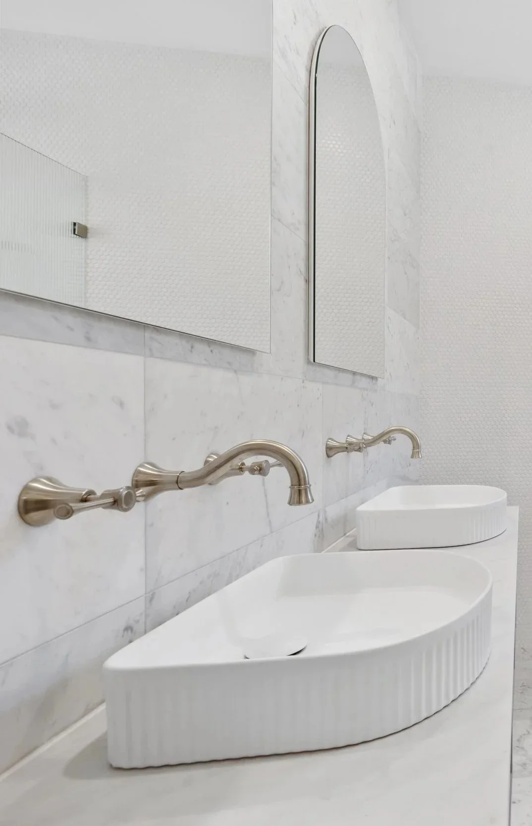A modern, minimalist bathroom with a white marble countertop, wall-mounted faucets, and a uniquely shaped white sink. A rectangular mirror and textured white wall tiles are visible.