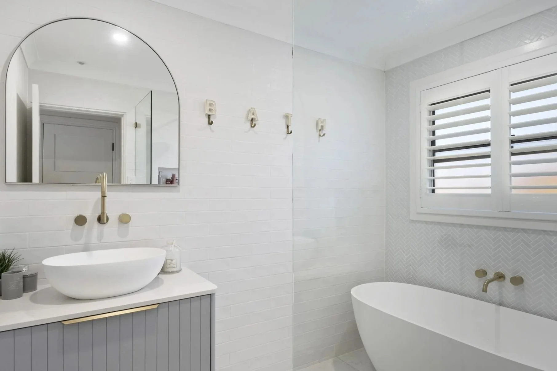 A modern, minimalist bathroom with white tiled walls, a white freestanding bathtub, a white vanity with a vessel sink, a large mirror, and a window with white shutters. Decor includes the word 'BATH' spelled out on the wall.
