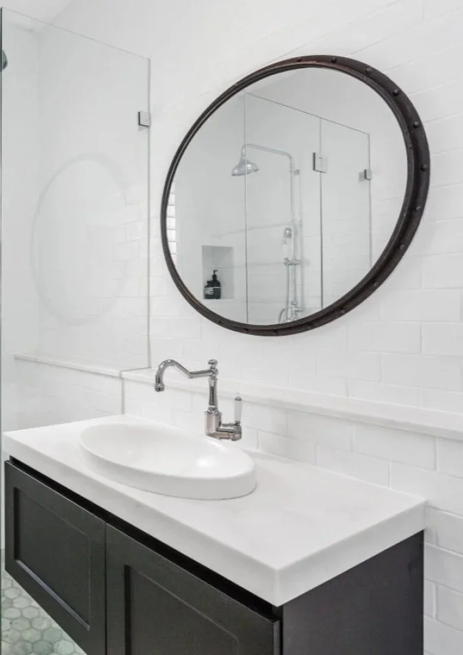 Modern bathroom with white brick wall, black-framed round mirror, white countertop sink, and a black vanity cabinet.