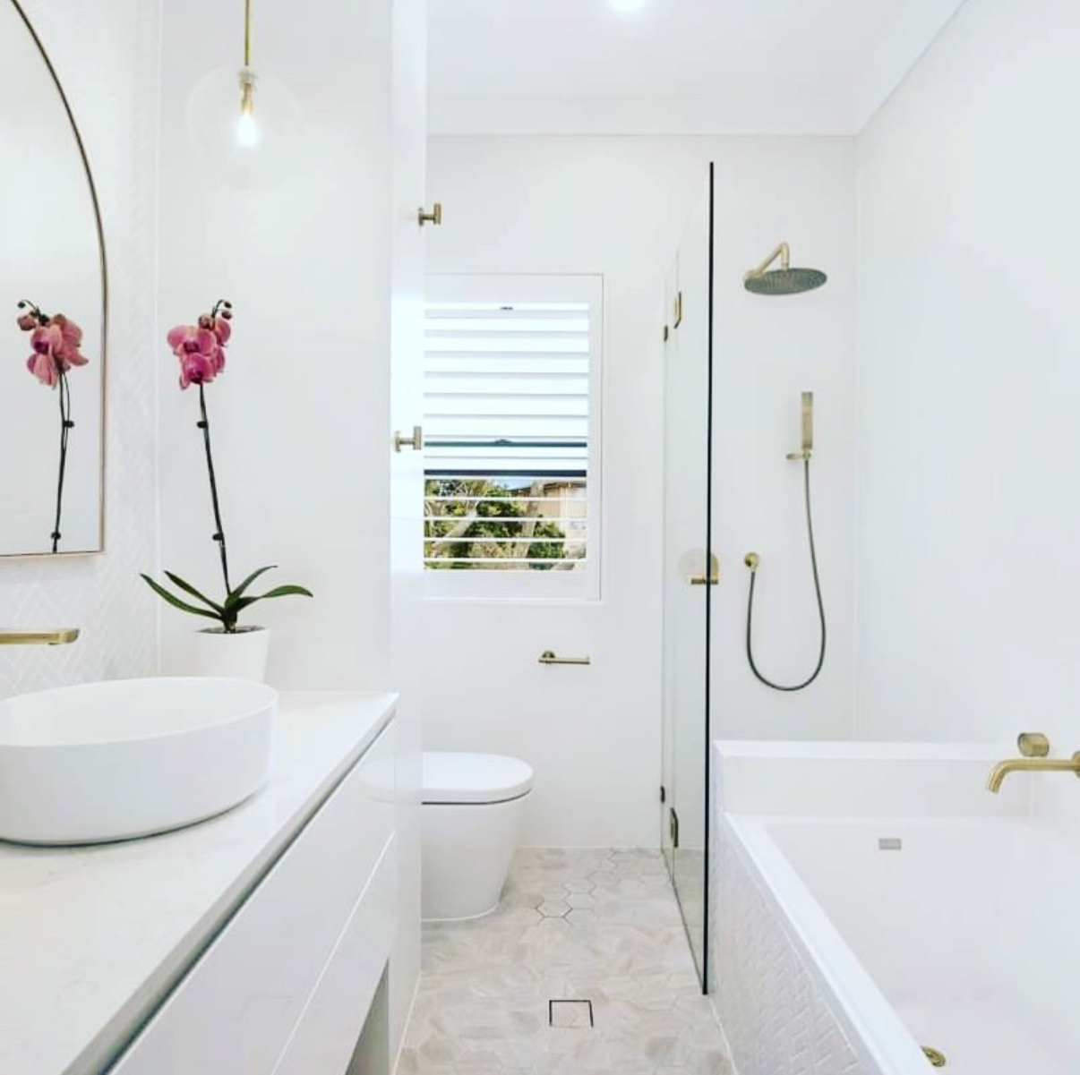 A modern white bathroom with a large oval vessel sink, a potted pink orchid, a gold light bulb fixture, a window with blinds, a toilet, a bathtub with gold fixtures, and a walk-in shower with a rainfall showerhead and handheld shower on the wall.