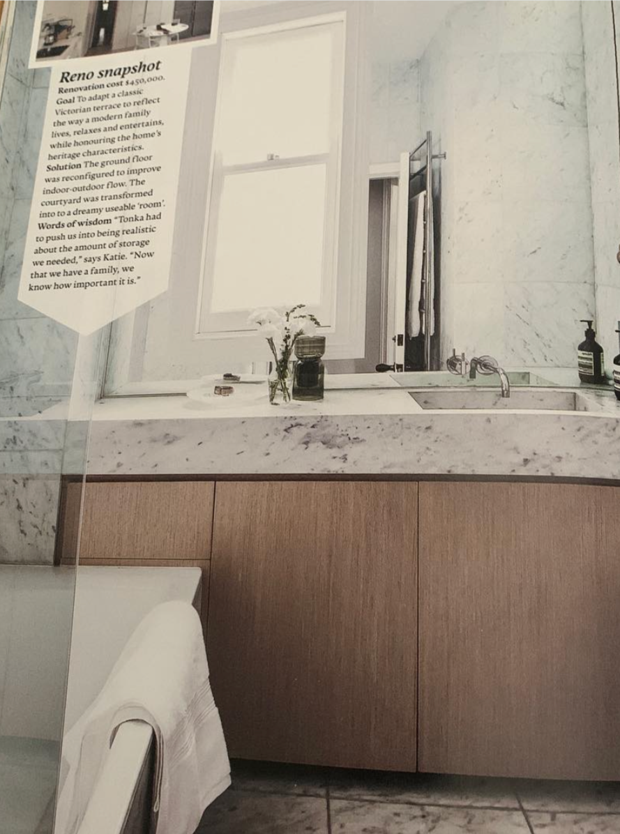 A bathroom with a marble countertop, a sink with a bronze faucet, a mirror, a window, and a black soap bottle on the right side of the counter. There are also some small decorative items and a small plant in a glass vase.