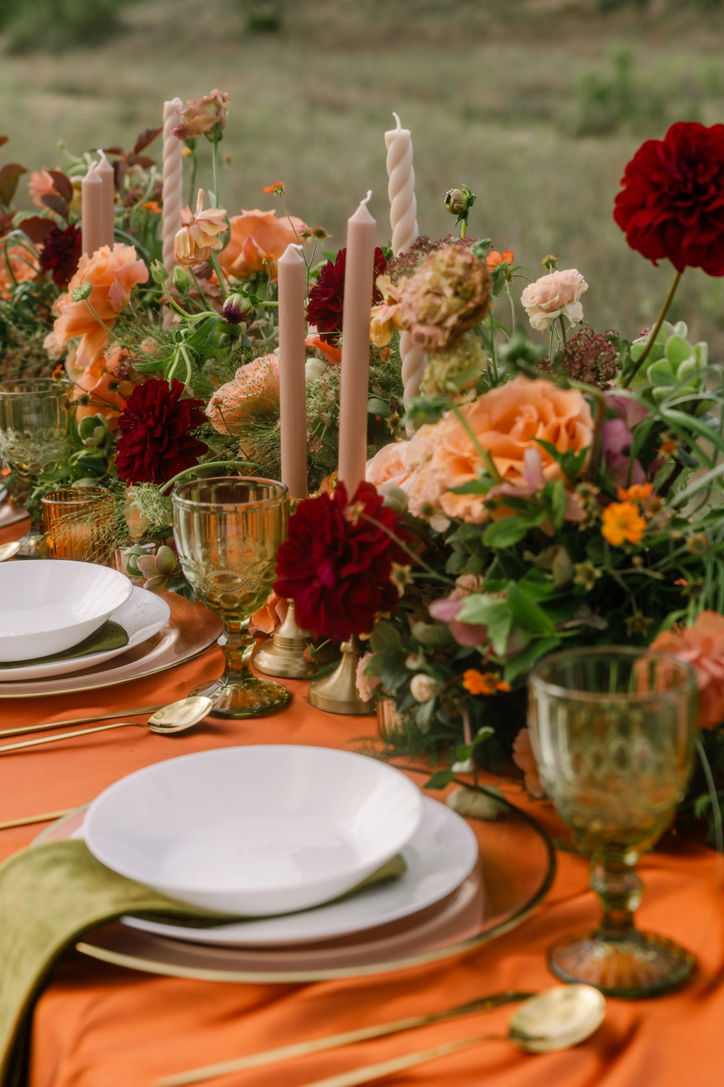 Decorative table setting with white plates, gold flatware, amber glassware, peach-colored tablecloth, and a floral centerpiece with various mixed flowers and candles.