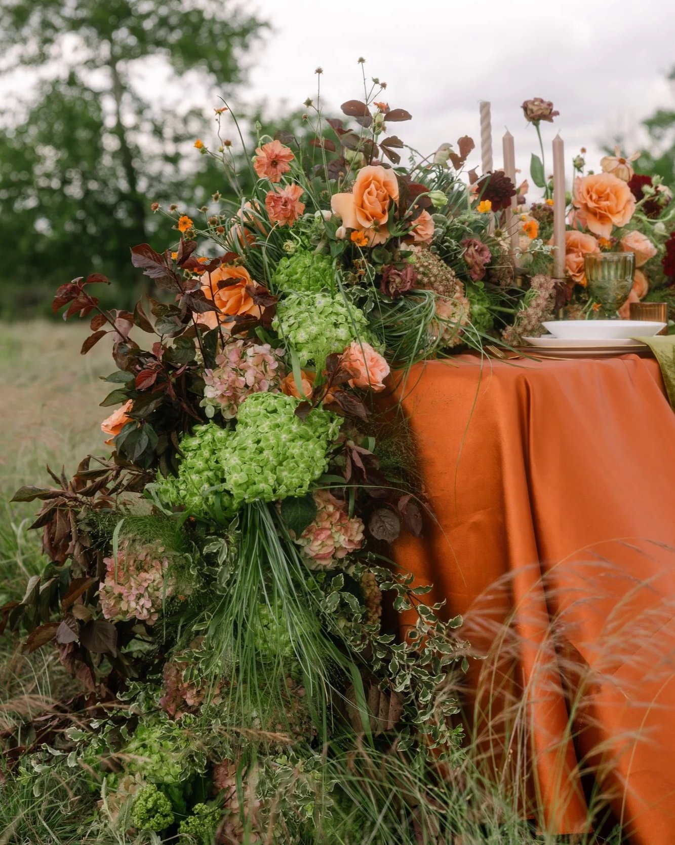 A table decorated with a large floral arrangement of pink, green, and burgundy flowers and greenery, draped with an orange tablecloth, outdoors with trees in the background.