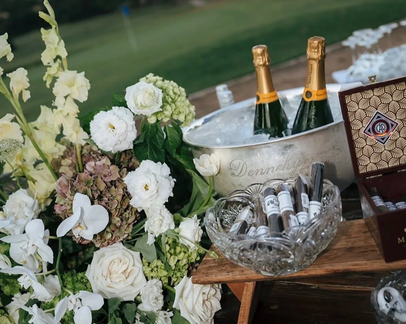 A table setup with white flowers, two bottles of champagne in an ice bucket, a glass bowl filled with rolled cigars, and a box of cigars, with a grassy area in the background.