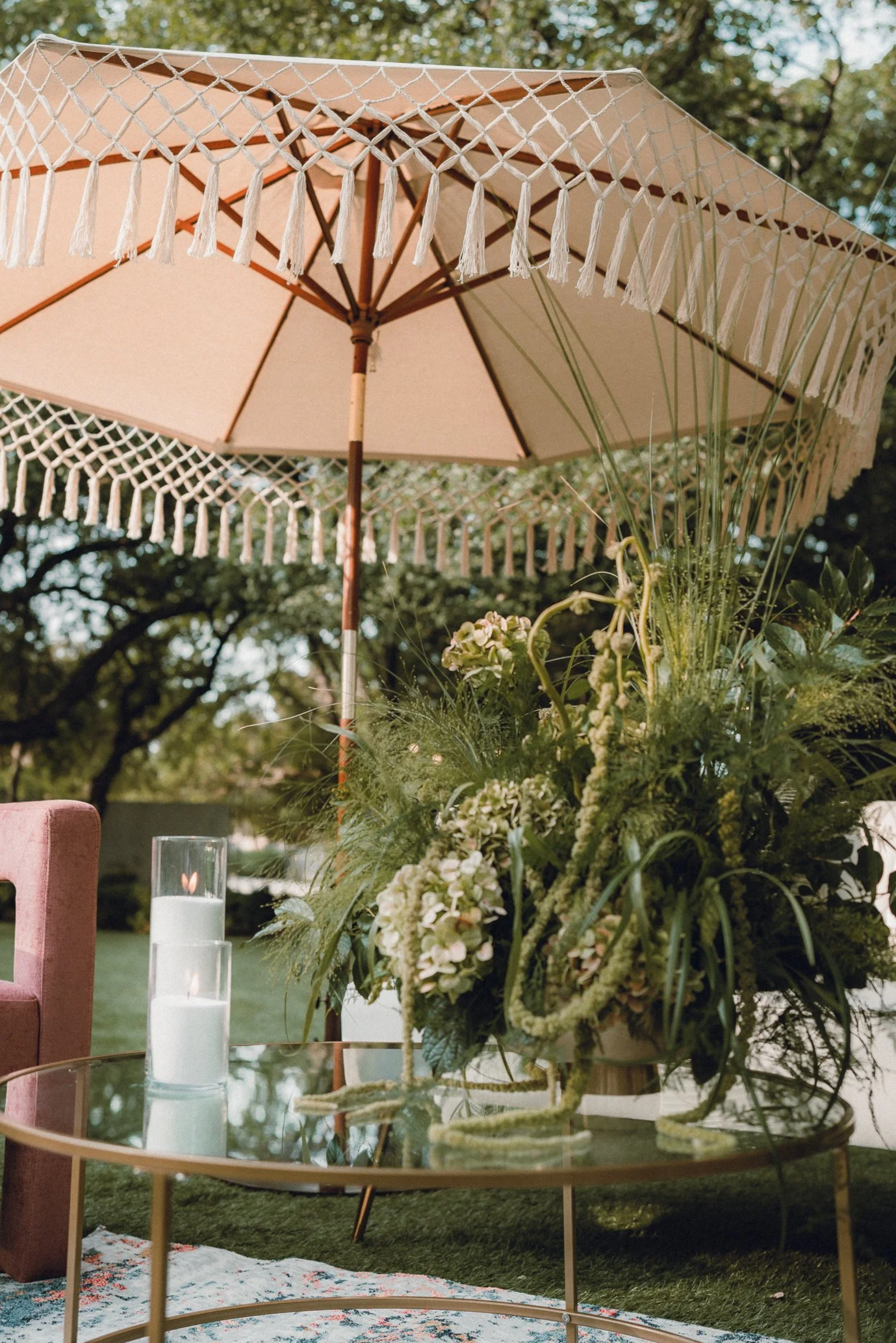A beige patio umbrella with decorative fringe and tassels over a floral arrangement of white and green flowers and foliage. In front of the arrangement, there is a glass table with a white candle inside a glass holder and a pink armchair visible on the left. The scene is outdoors with trees in the background.