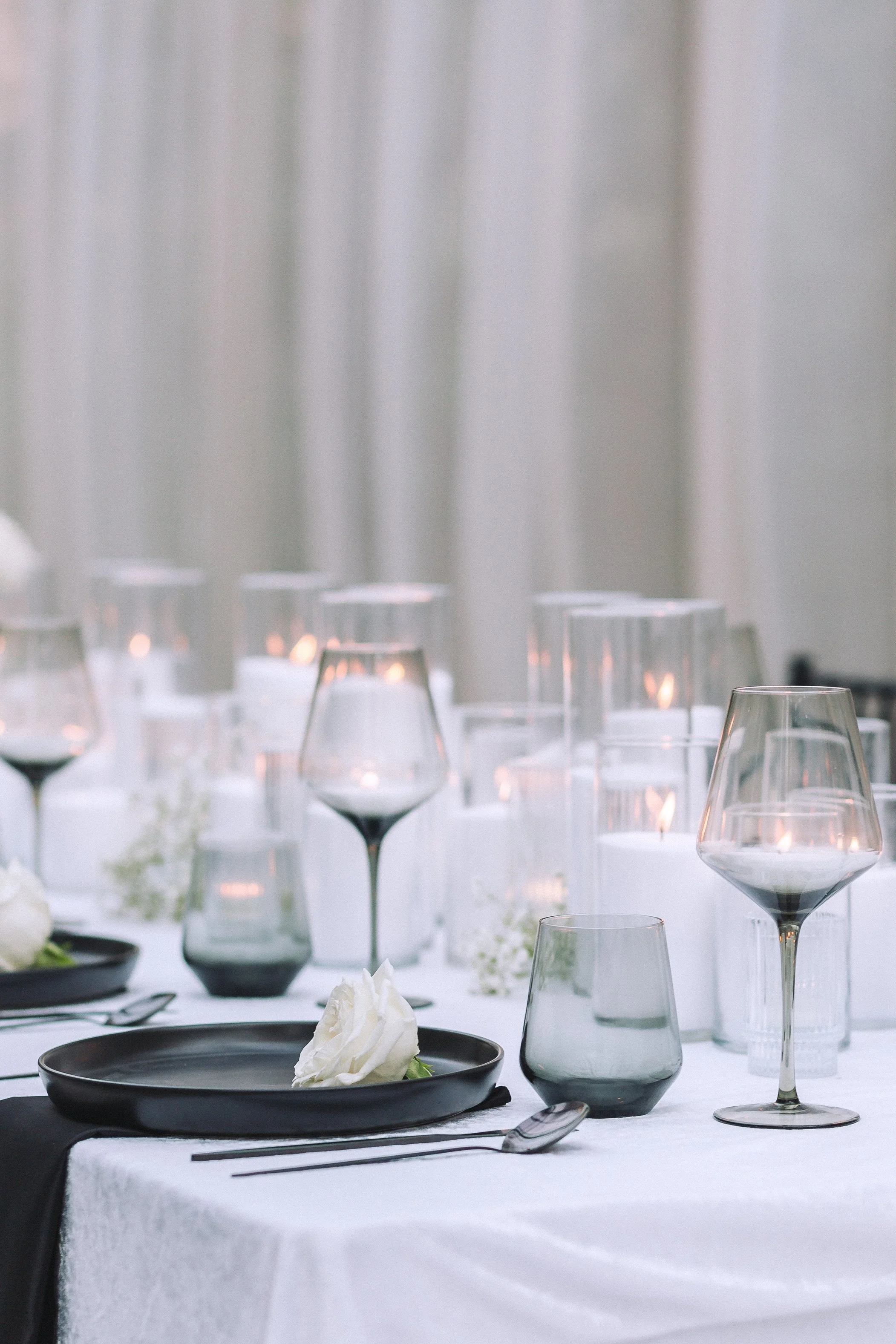 Elegant table setting with wine glasses, plates, and a black napkin on a white tablecloth, with a blurred background of white curtains.