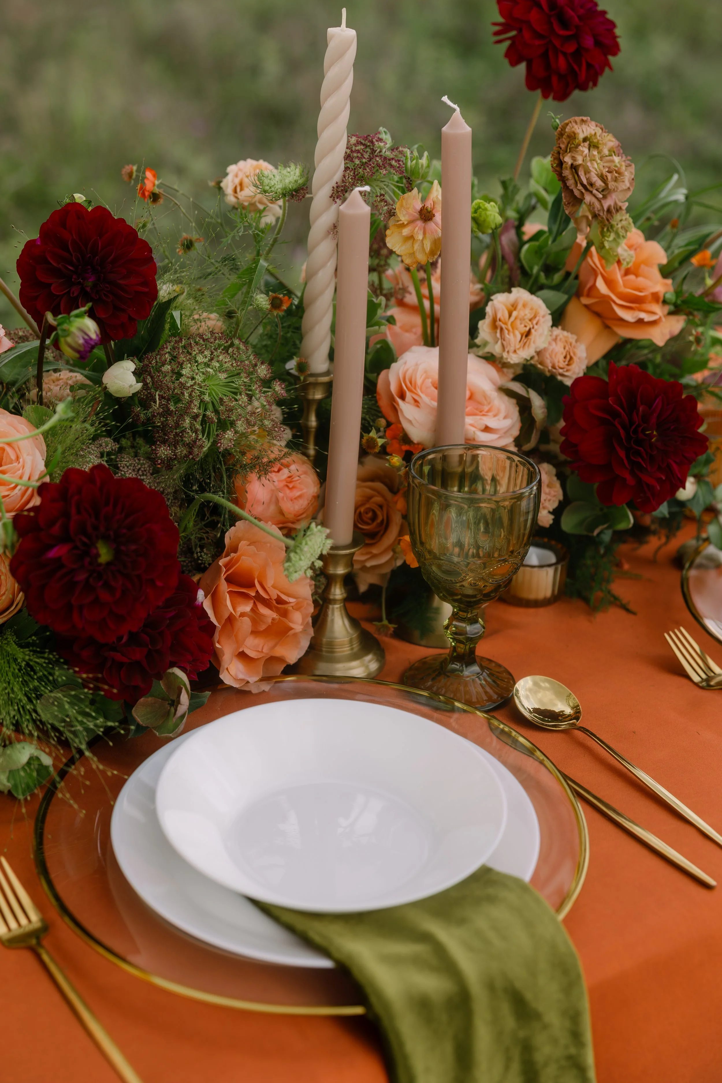 A table setting with a white plate and gold-rimmed glass, gold utensils, surrounded by a floral centerpiece with red and peach flowers, and three white candles, on a rust-colored tablecloth.