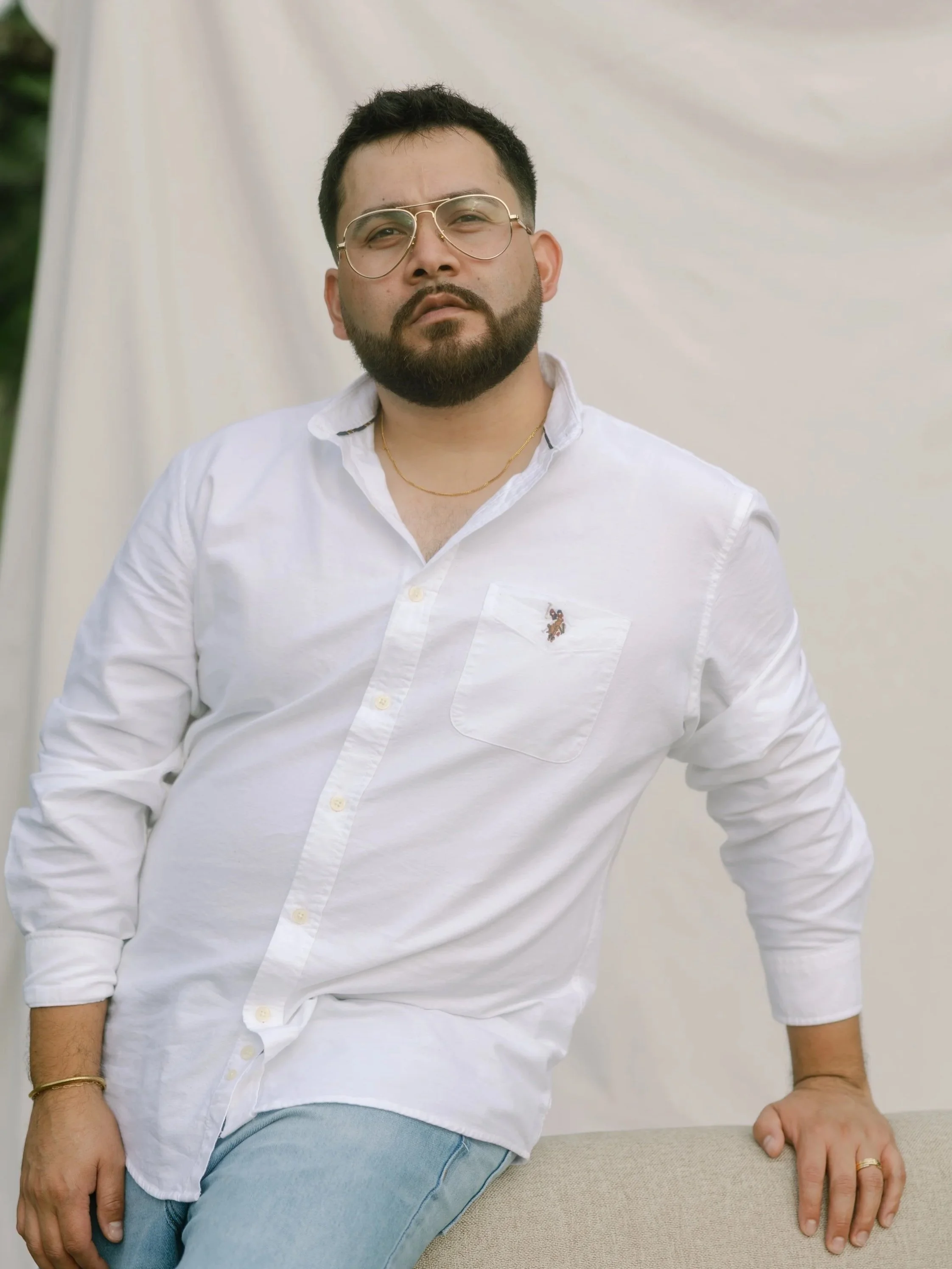 A man with dark hair and glasses, wearing a white shirt and light blue jeans, standing against a neutral background.
