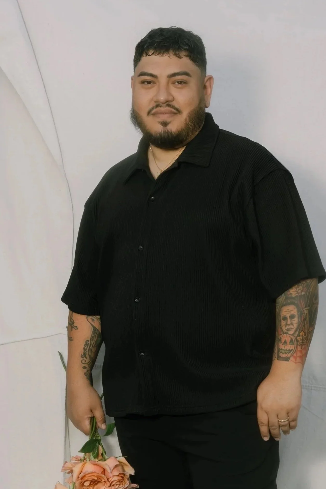 A man with dark hair, beard, and tattoos, wearing a black short-sleeve button-up shirt, holding pink roses, standing against a plain white wall.
