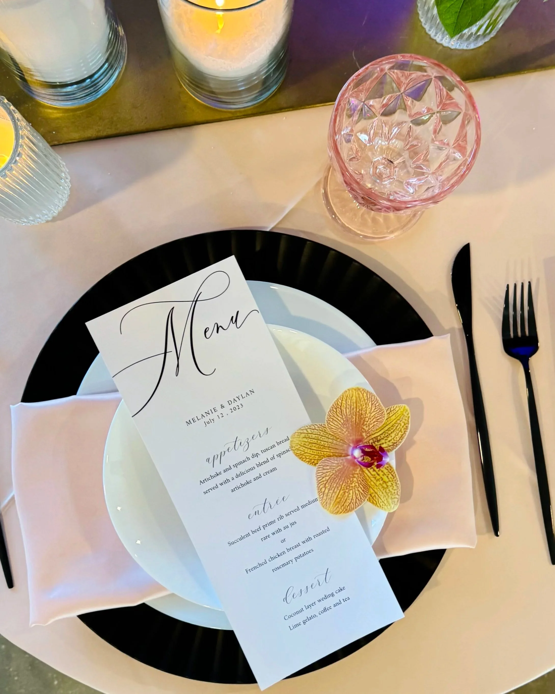Elegant place setting for a wedding event with a menu card, a black plate, a pink glass, and three candles in glass holders, alongside a white tablecloth.