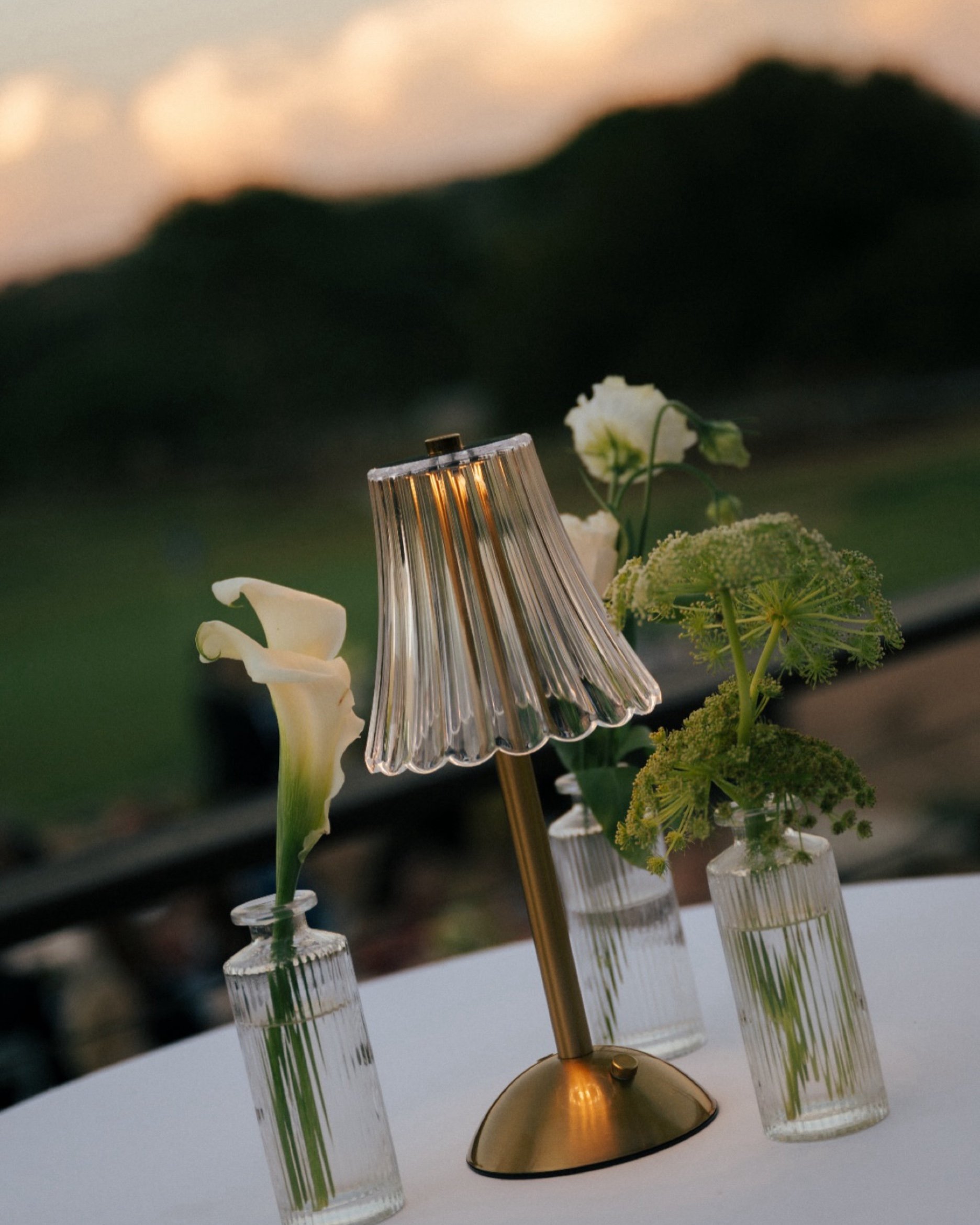 Table centerpiece with a glass lamp and flowers in small glass vases against a blurred background.