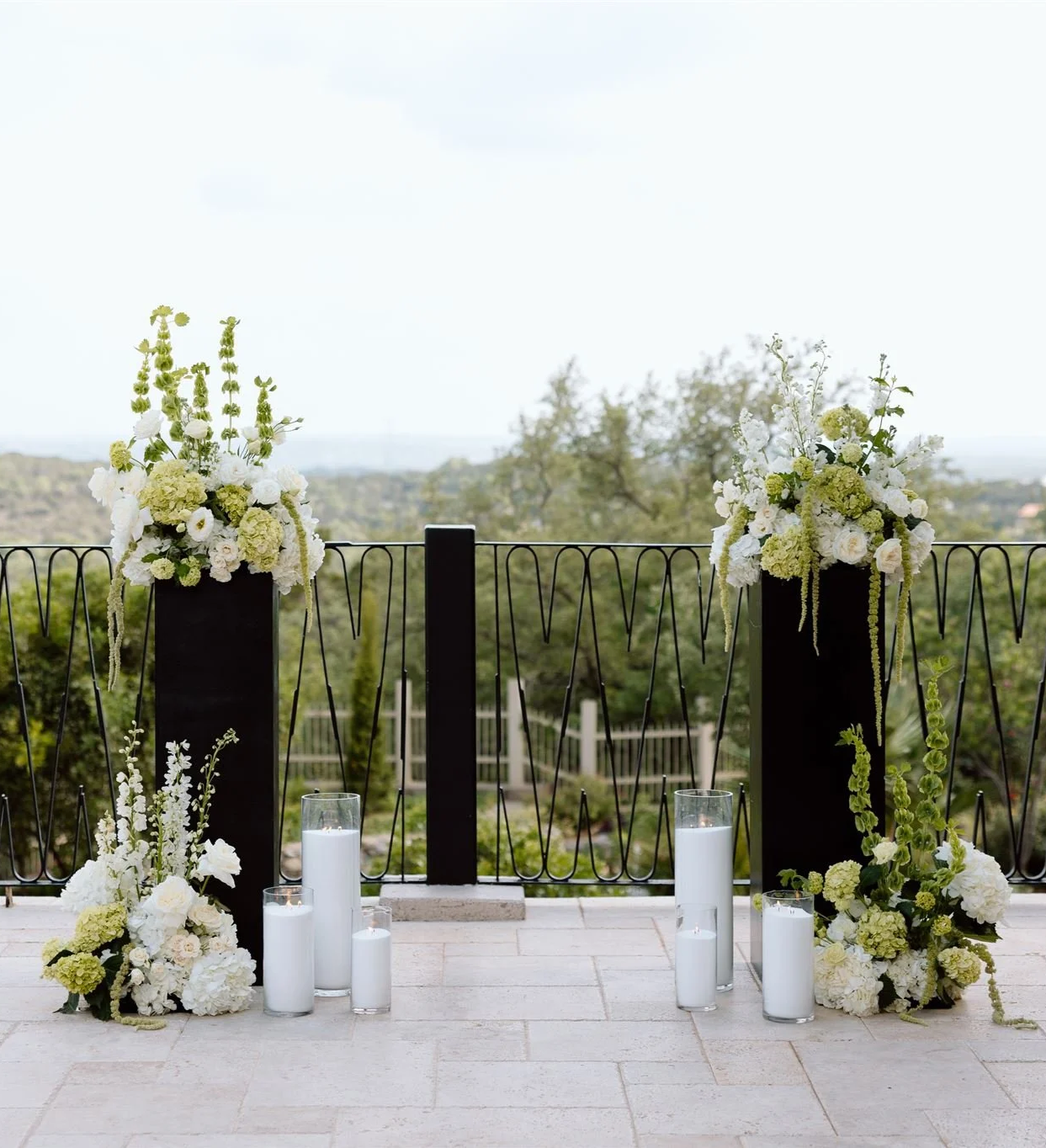 Beautiful outdoor wedding or event setup with black floral arrangements and white candles on a stone patio, overlooking a scenic landscape with trees and cloudy sky.