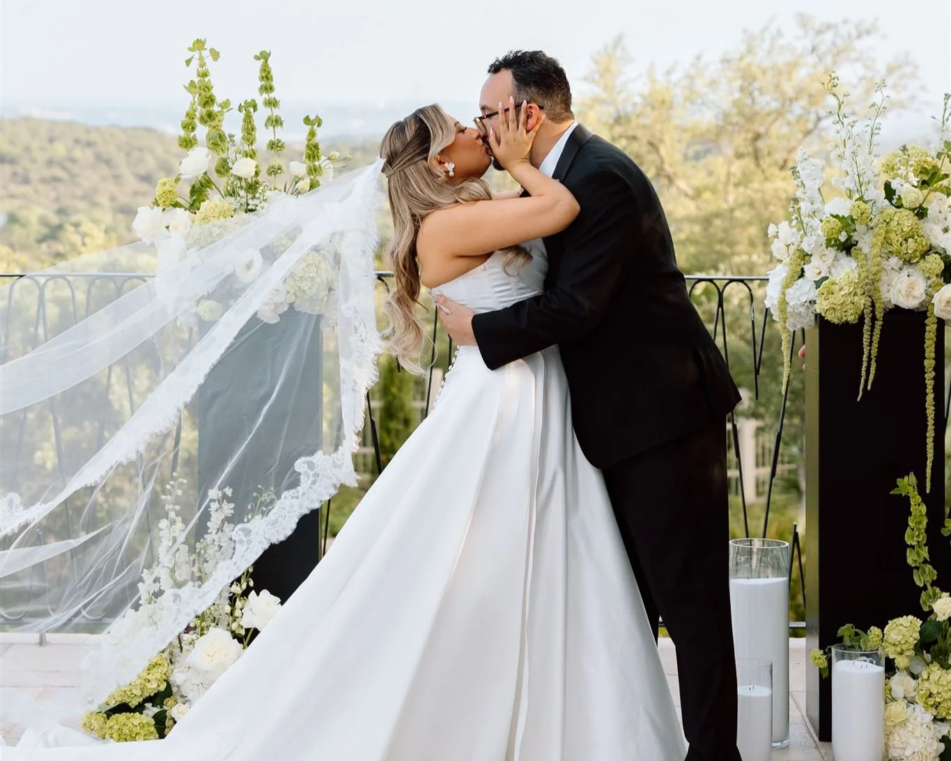 A bride and groom kissing at their outdoor wedding ceremony, with floral arrangements and candles around them.