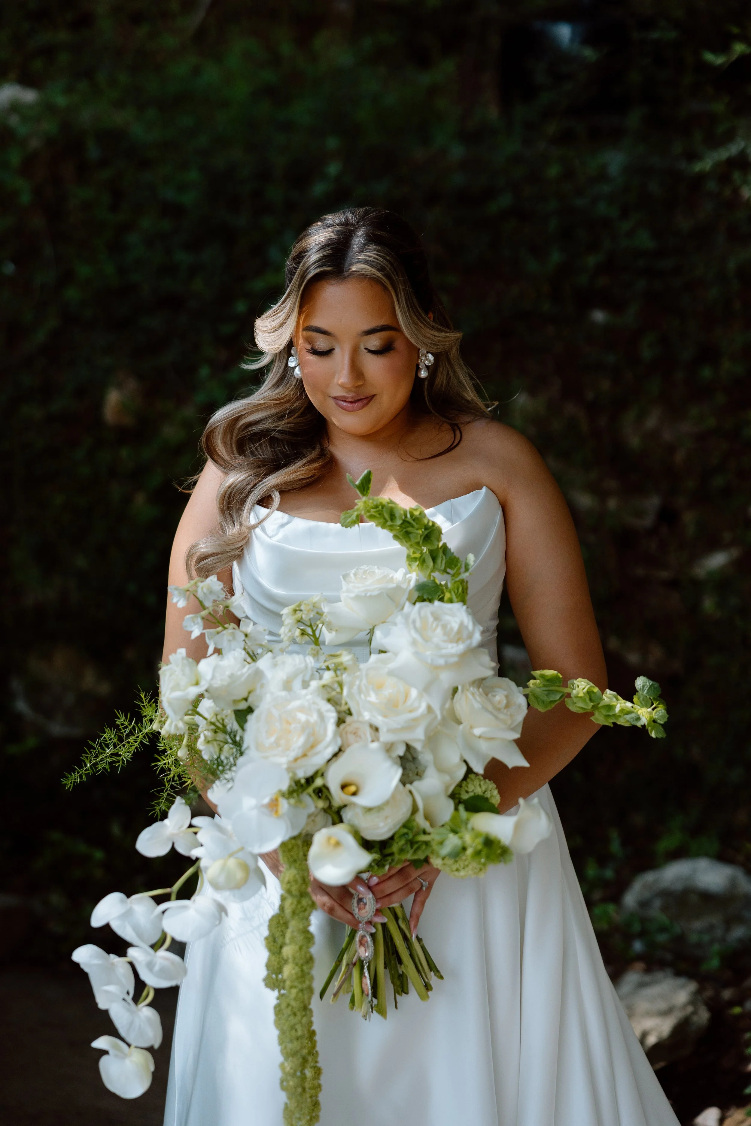 A woman in a white wedding dress holding a large bouquet of white flowers, standing outdoors with greenery in the background.