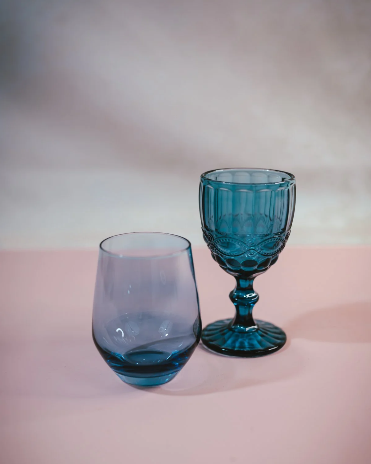 Two blue glass cups, one with a rounded shape and the other with a more decorative stem and intricate design, placed on a pink surface with a soft gradient background.