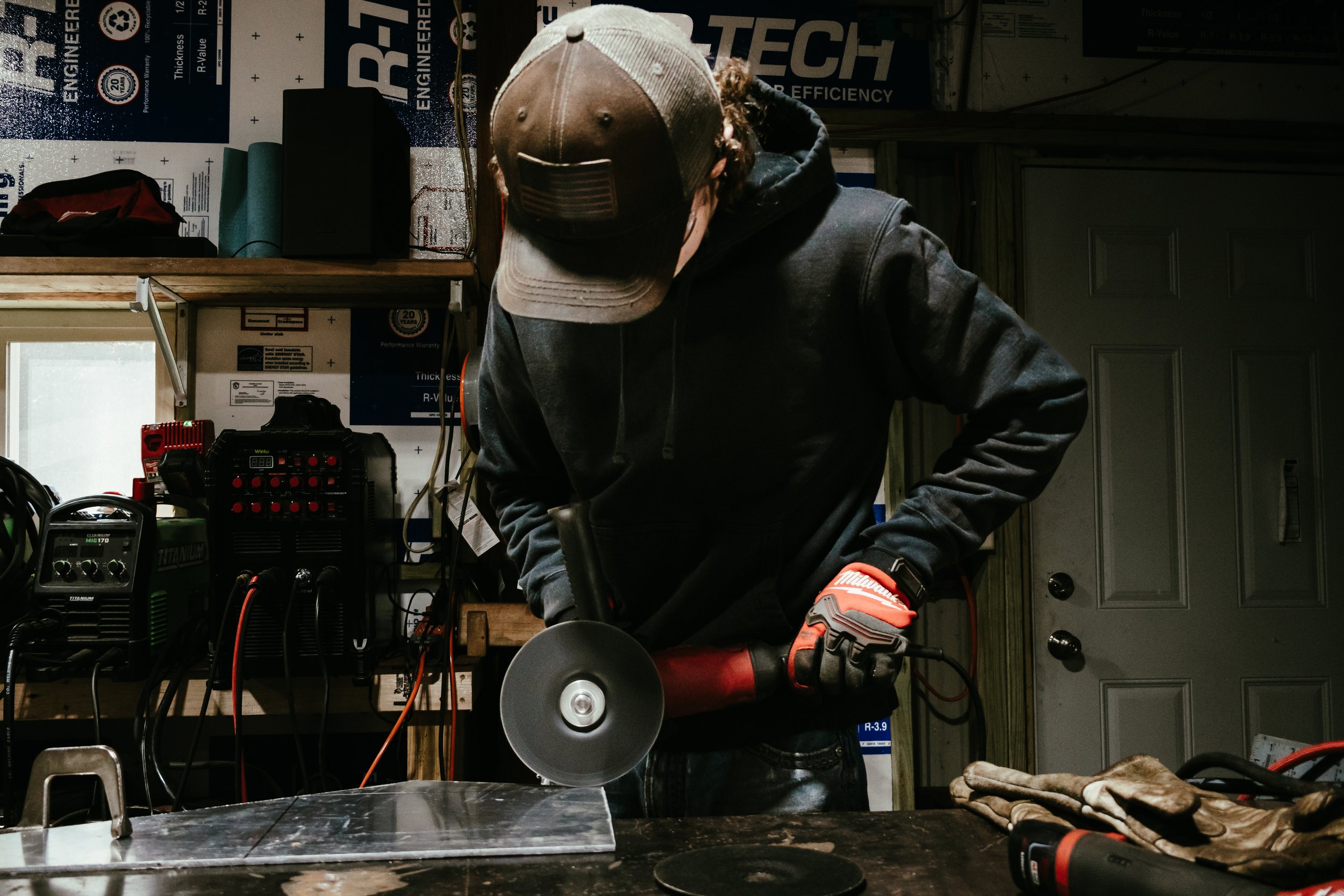 Man wearing a gray baseball cap, hoodie, and gloves using a power tool in a workshop with various tools and equipment in the background.