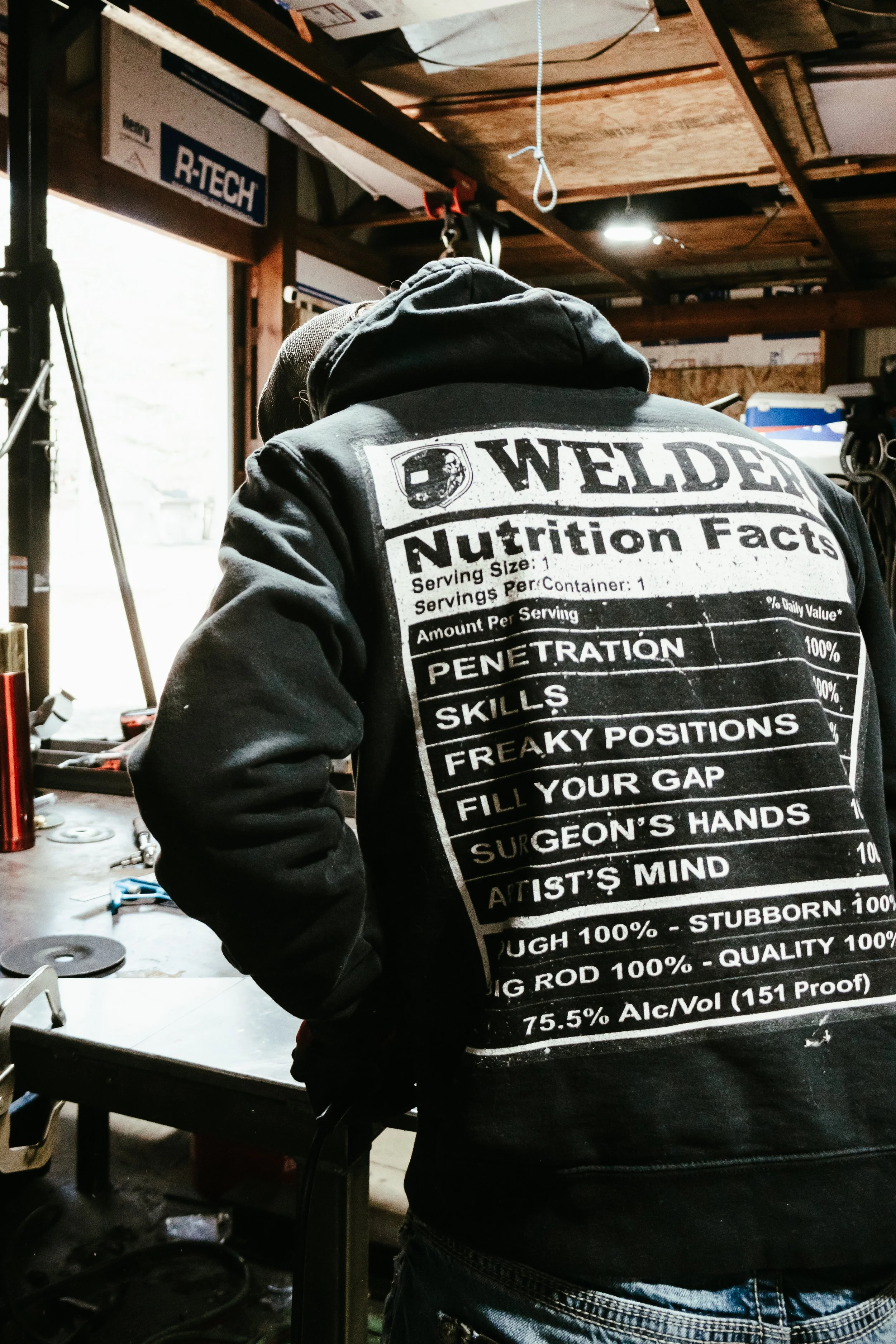 A person wearing a black hoodie with printed text on the back, standing at a workbench in a garage or workshop, with tools and equipment around.