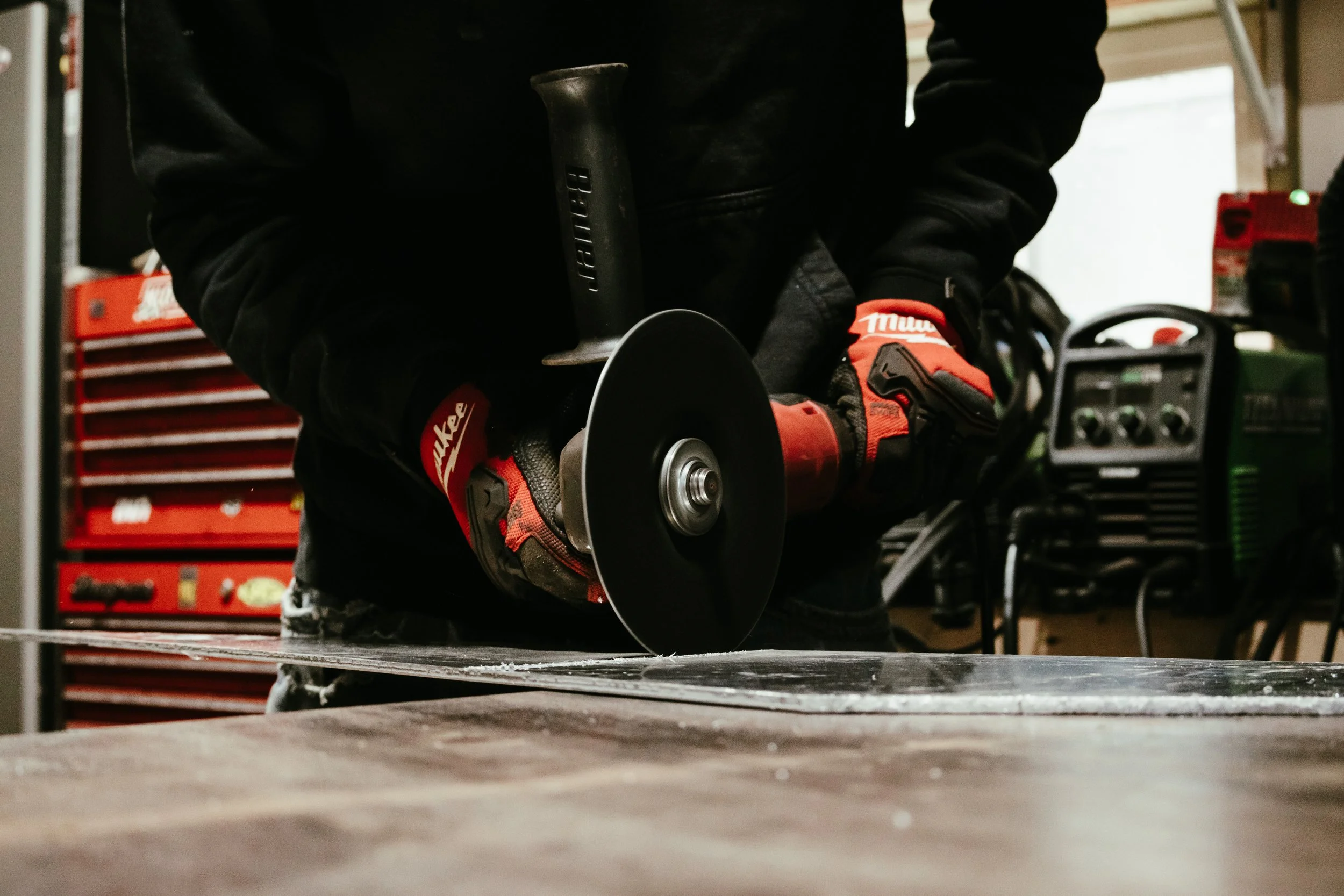 Person wearing red and black gloves using angle grinder on a metal surface in a workshop.