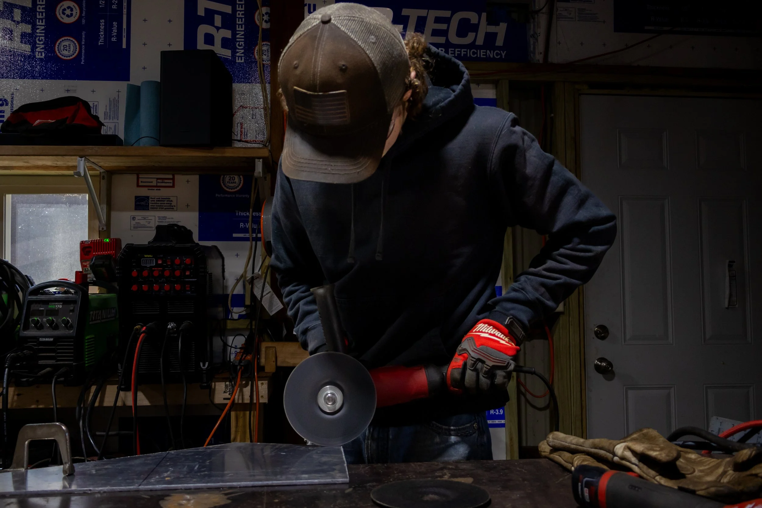 A person wearing a brown cap, black hoodie, and red gloves working with a power tool in a workshop with various tools and equipment around.