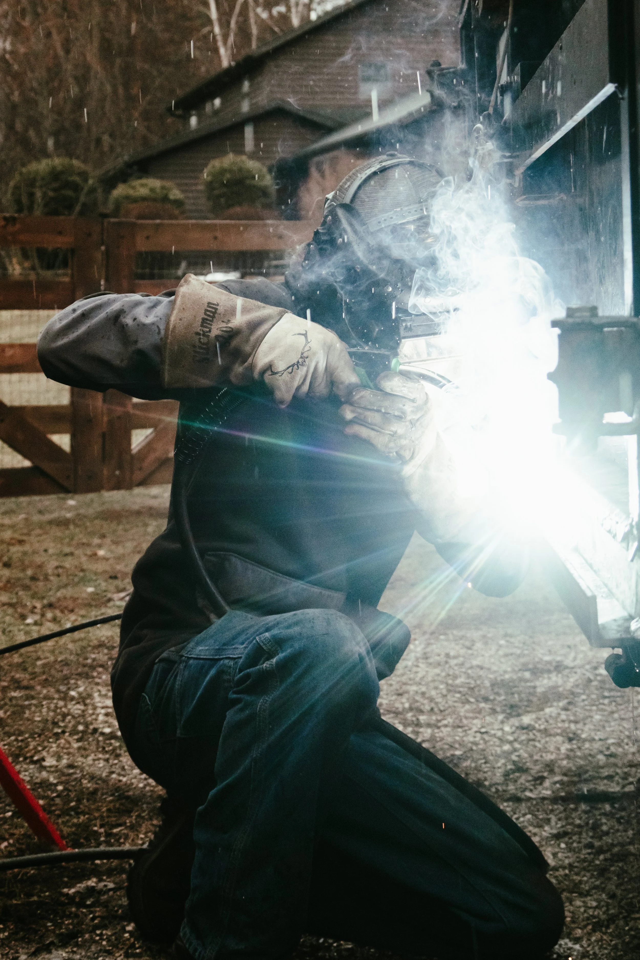 A person welding outdoors at night, wearing protective gear and kneeling on the ground, with sparks and bright light from the welding process visible.