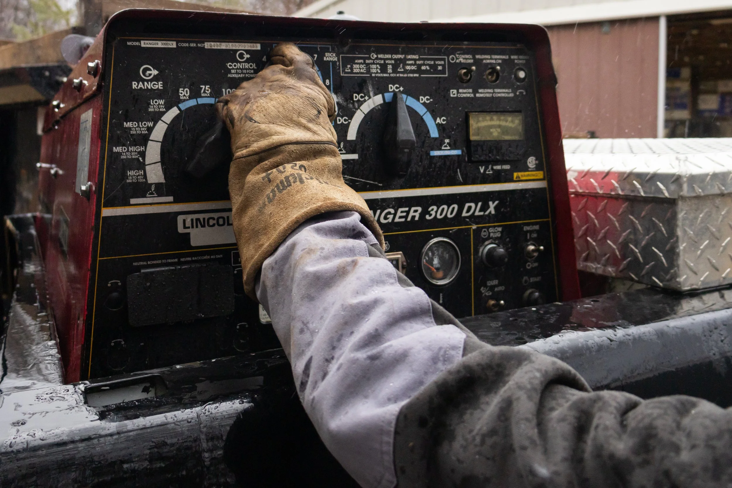 A gloved hand operating a Lincoln Ranger 300 DLX welding machine outdoors with trees and a shed in the background.