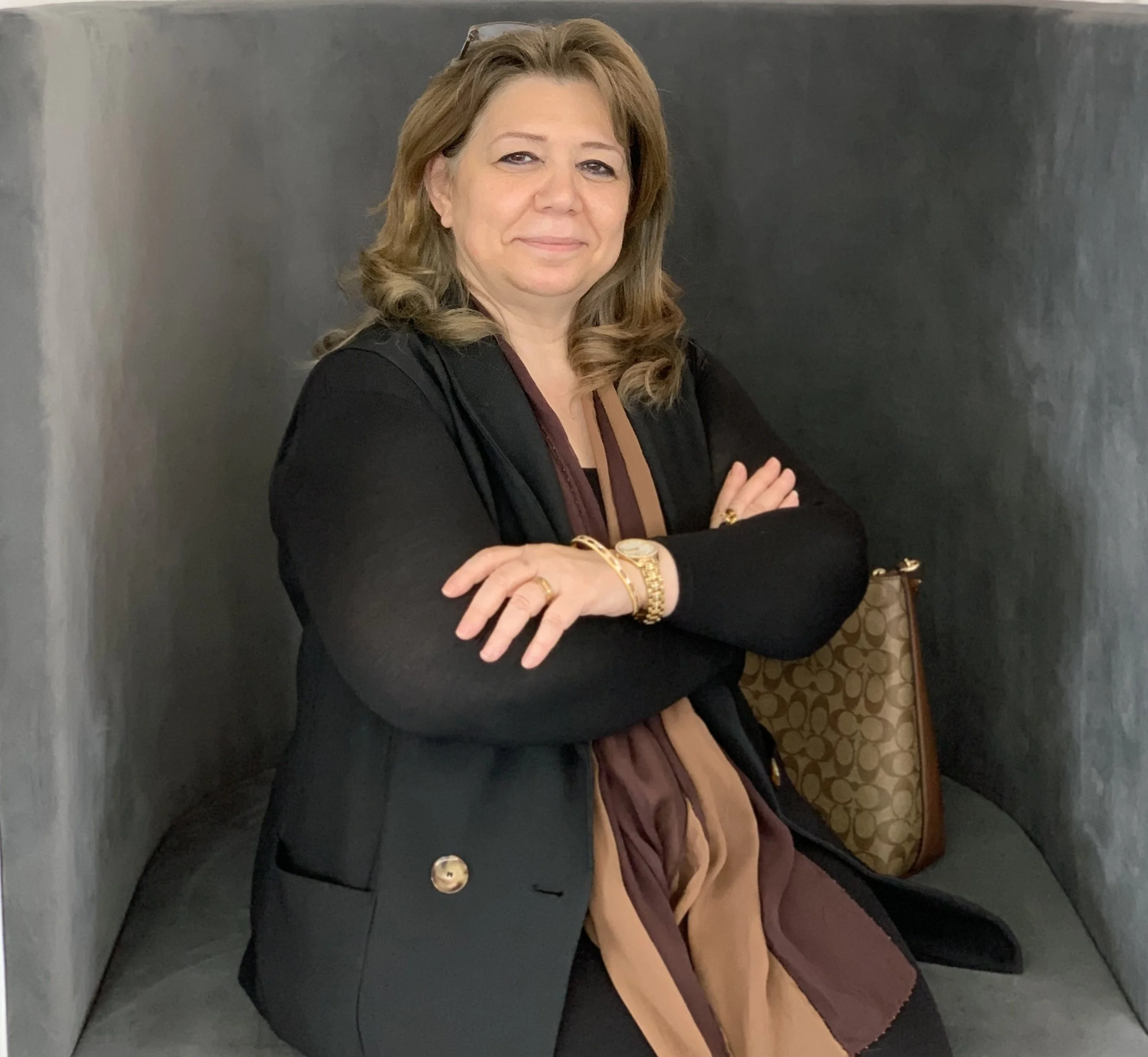 A woman with shoulder-length blonde hair, wearing a black blazer, sitting in a gray, semi-enclosed corner with her arms crossed, holding a brown Coach handbag with a gold pattern, and wearing gold jewelry.