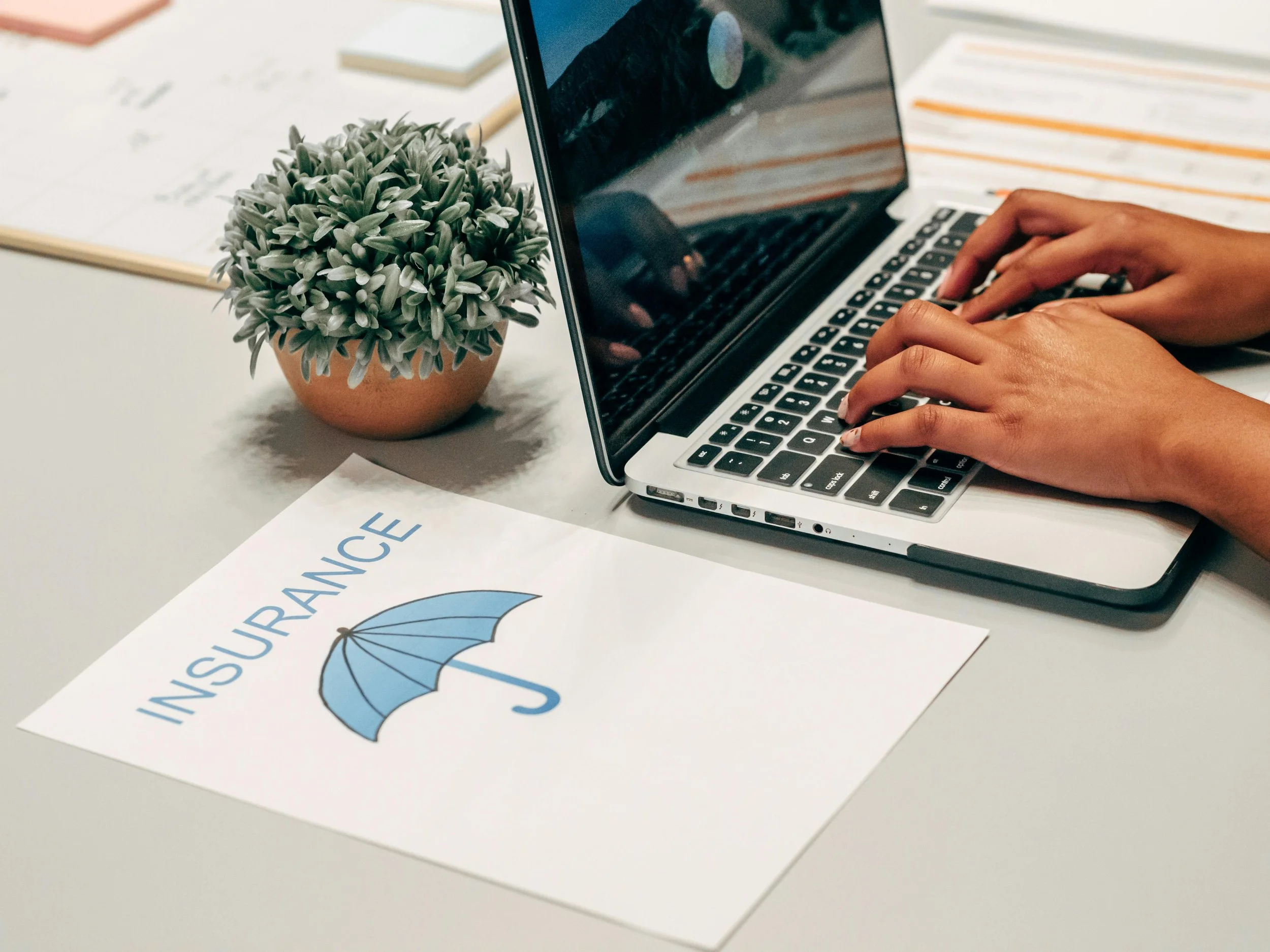 A person's hands typing on a laptop keyboard, next to a potted plant and insurance paperwork with an umbrella graphic on a desk.