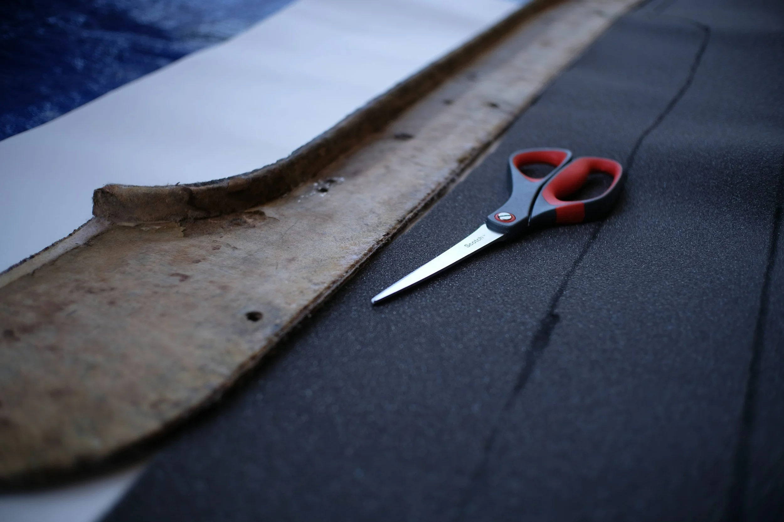 A pair of scissors with red and black handles lying on a dark surface near a buildup of old, peeling adhesive on a flooring edge.