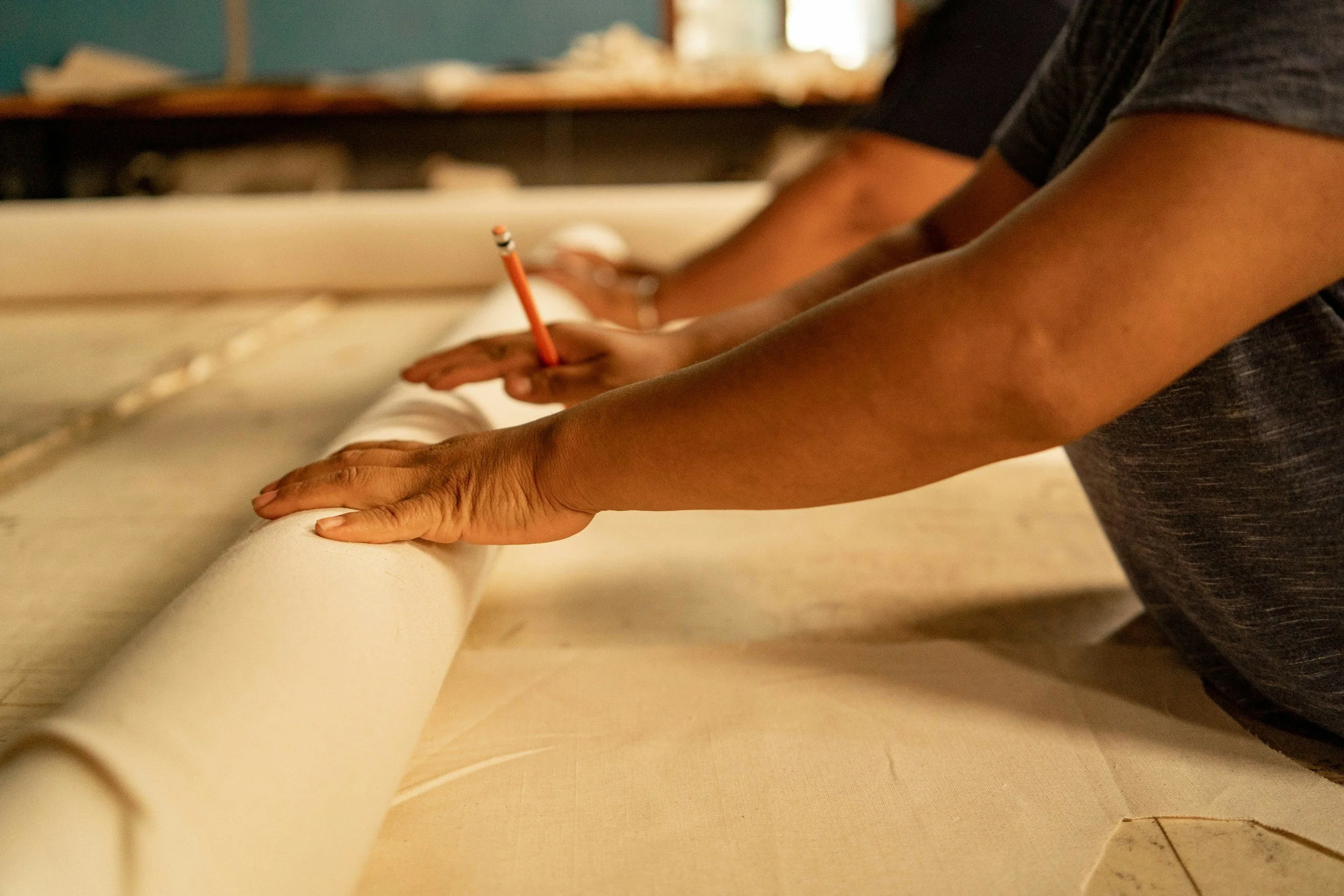 A person holding a pencil, writing or drawing on a piece of paper or canvas. Only their arms and hands are visible.