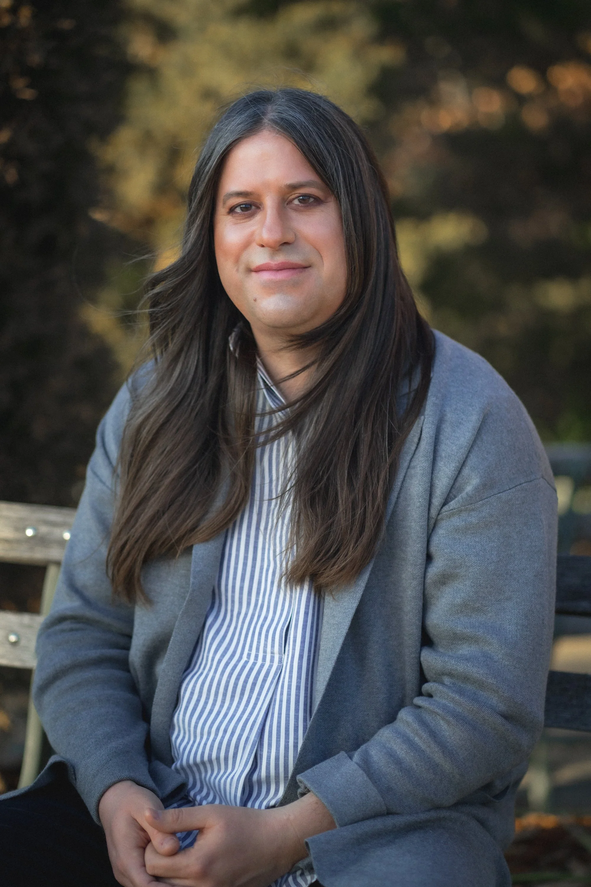 A person with long dark hair, wearing a gray blazer and a blue and white striped shirt, sitting outdoors with a blurred background of trees, looking directly at the camera.