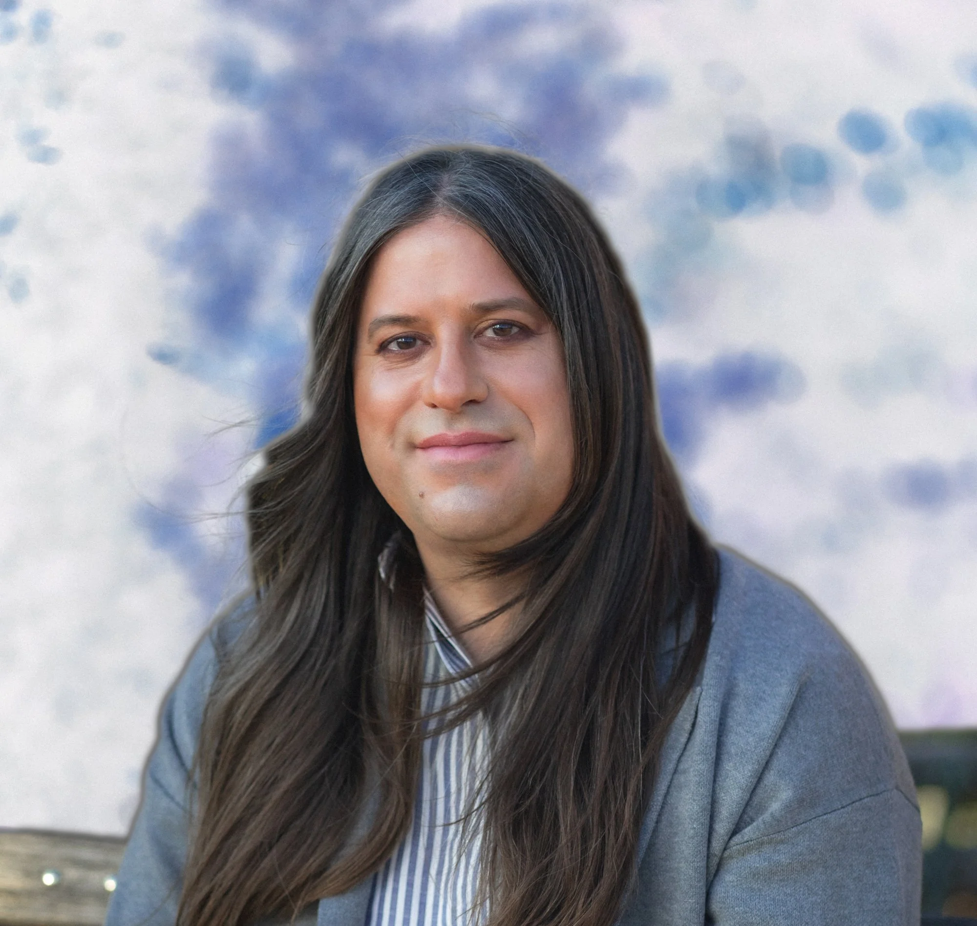 Close-up of a woman with long dark hair, wearing a gray sweater and a light blue and white striped shirt, outdoors with a cloudy sky in the background.