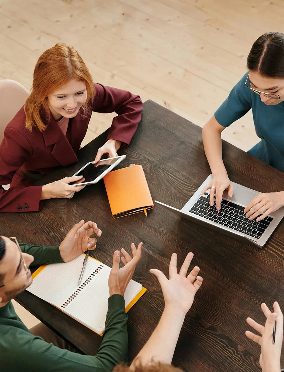 Group of four people having a meeting at a dark wooden table, with one woman using a laptop, another holding a tablet, and two others gesturing with hands, with notebooks and pens on the table.